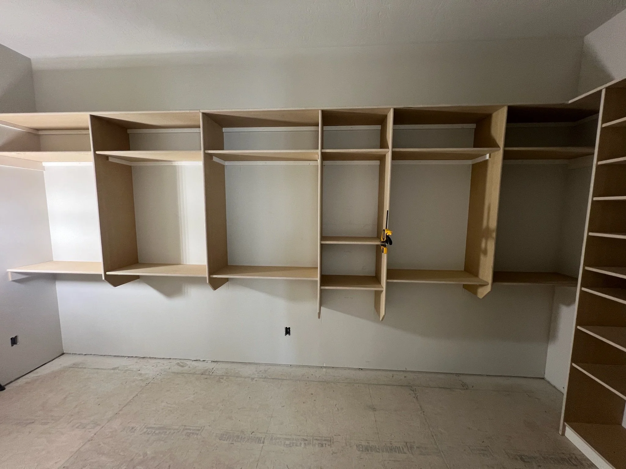 Empty wooden closet or shelving unit with multiple compartments and shelves, located in a room with unfinished flooring and white walls.