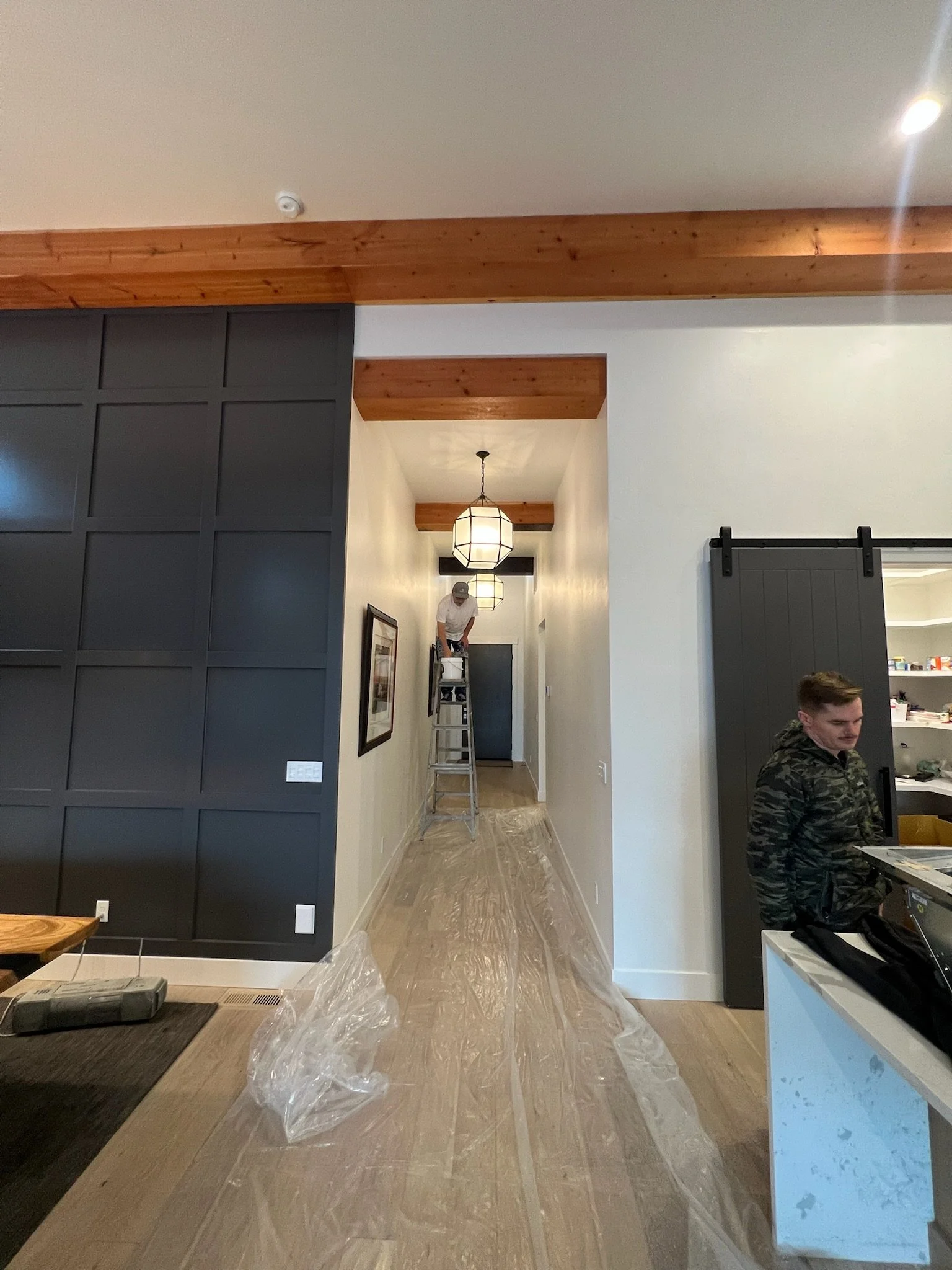 A man standing on a ladder in a hallway, with two large hanging light fixtures and a wood ceiling beam. Two young men are sitting in the foreground, one at a table and the other at a counter, in a modern interior space.