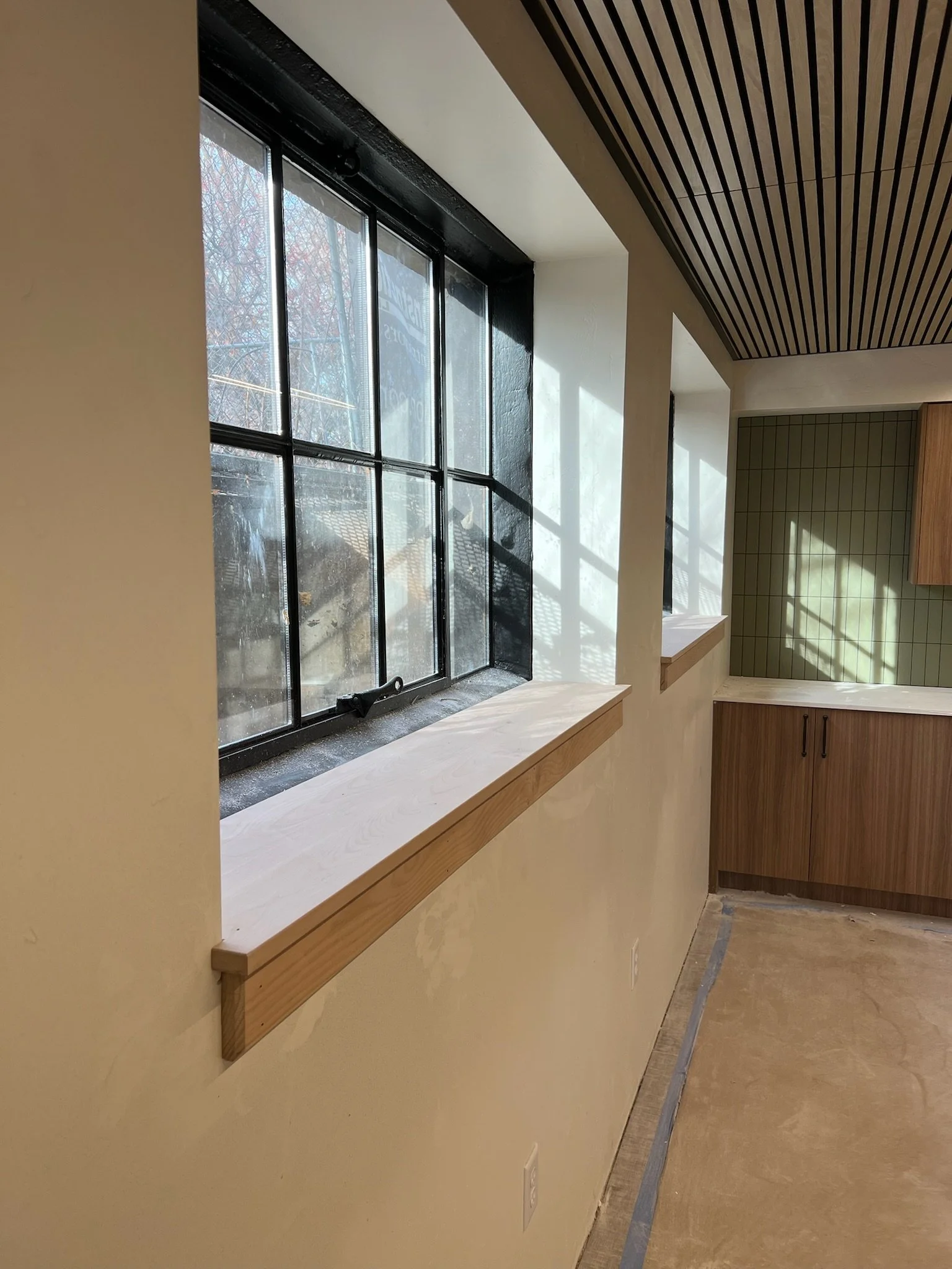 Photo of a window with black metal framing and a view of outdoor trees. The window is set into a light beige wall with a wooden ledge underneath. The ceiling has black and wood striped paneling, and there are green tiled walls and wooden cabinets in 
