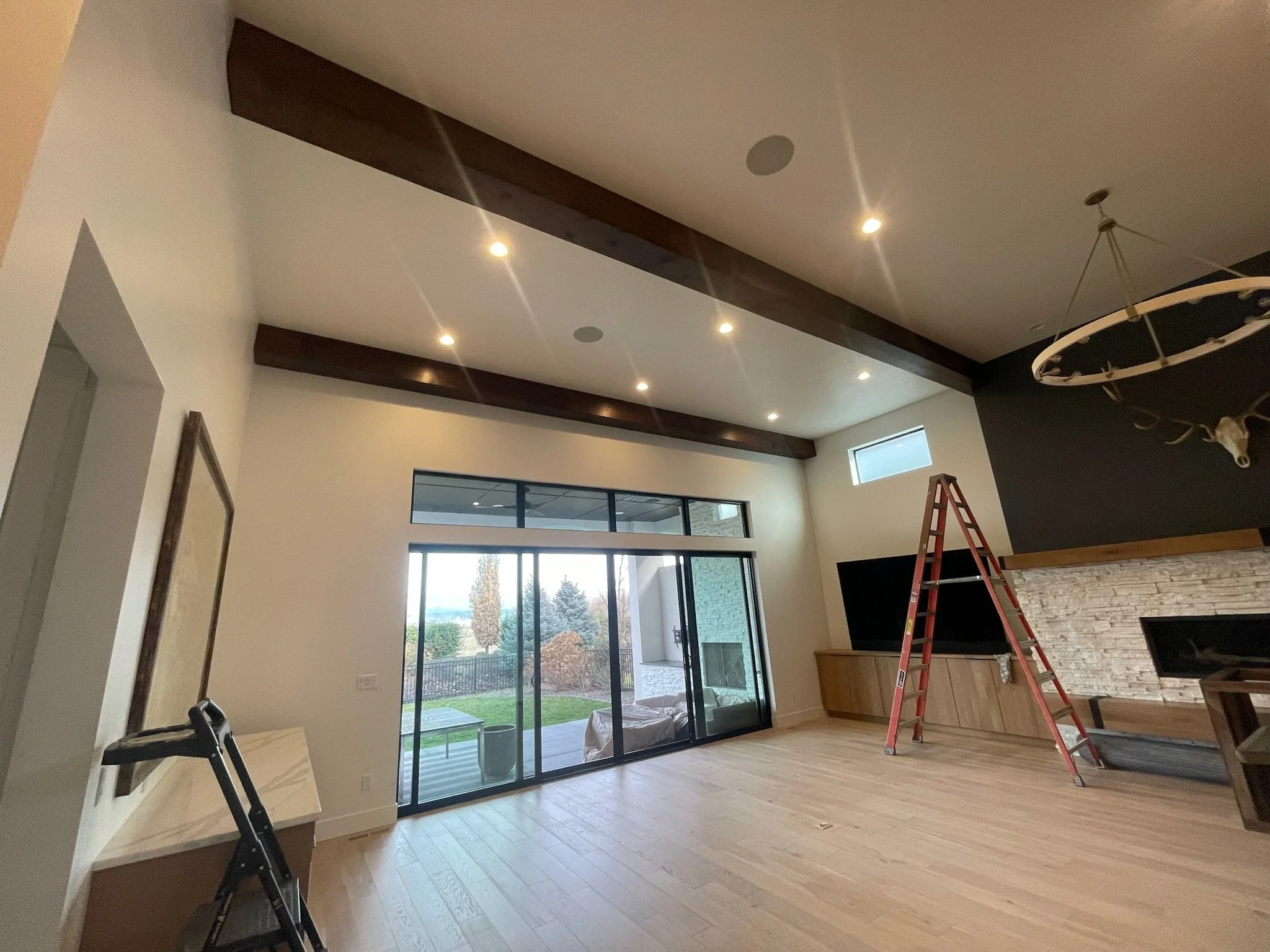Interior of a modern living room with high ceiling, dark wooden beams, a large glass sliding door leading to a backyard, and a fireplace with a stone surround. There is a ladder, a black chair, and a deer head mounted on the dark wall.
