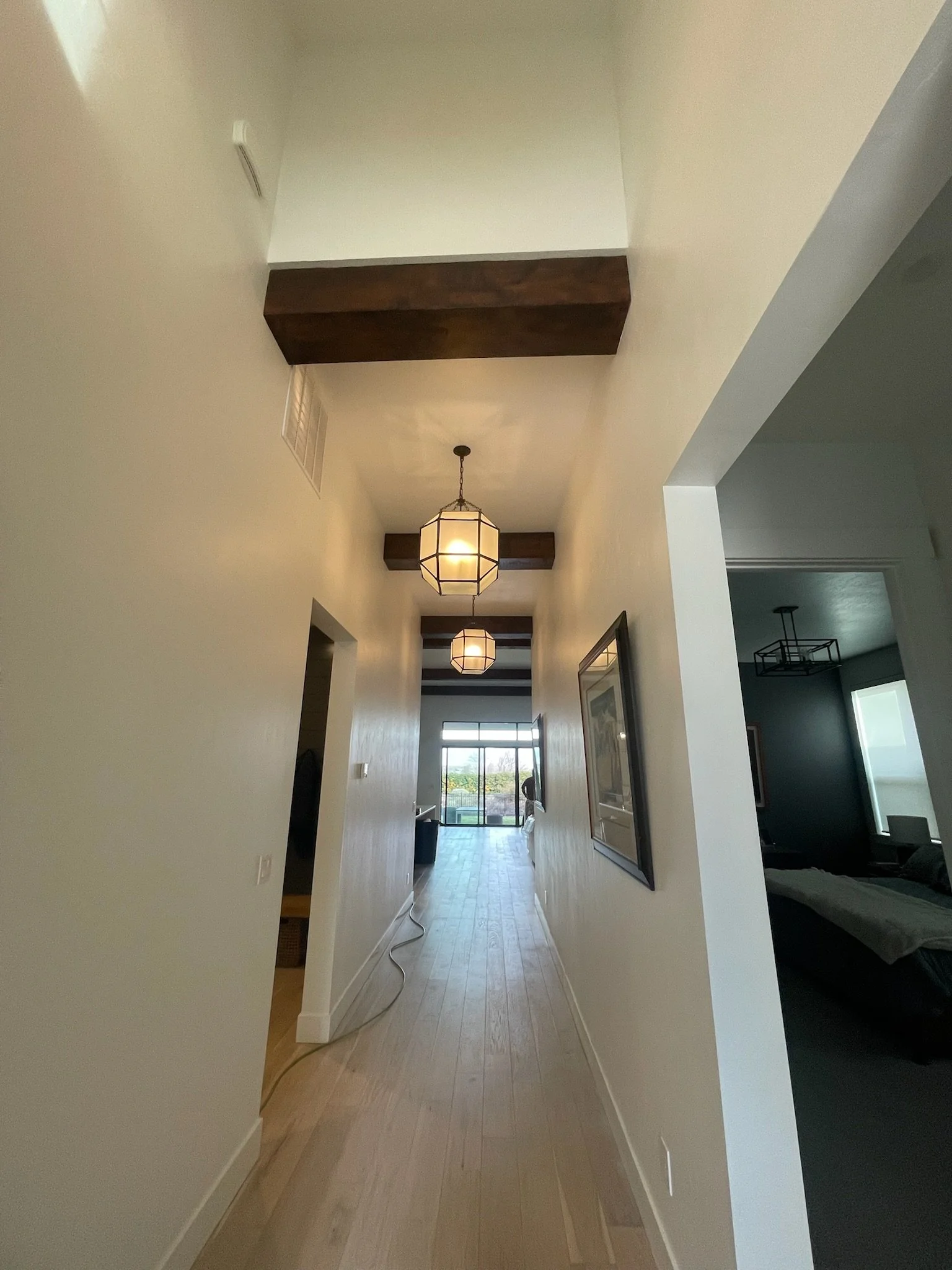A hallway with light-colored wooden flooring, white walls, hanging light fixtures, and a large window at the far end showing an outdoor scene. There are dark wood beams on the ceiling and some framed pictures on the right wall.