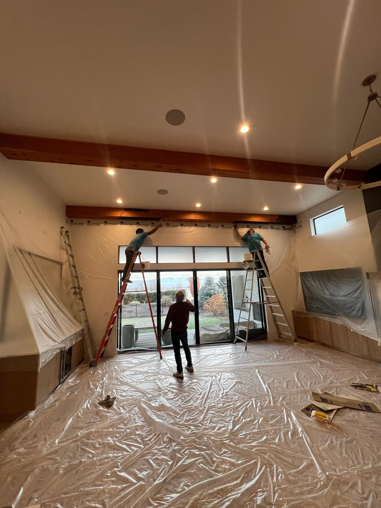 Two workers on ladders install or paint a high ceiling in a room, with plastic sheeting covering furniture and flooring for protection.