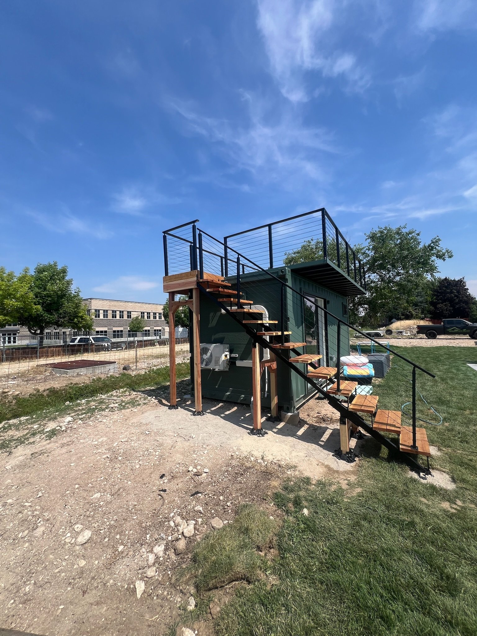 A small green building with a staircase leading to a rooftop deck with metal railing, outdoors on a sunny day, trees and a parking lot in the background.