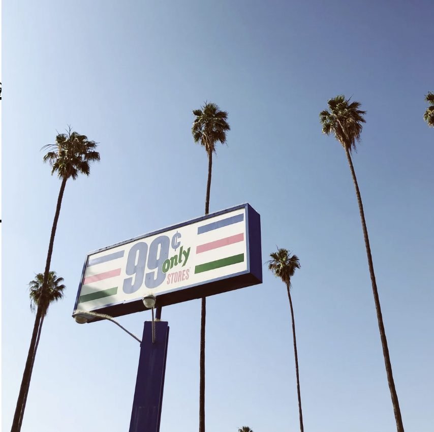 A sign for 99-cent stores under a clear blue sky, with tall palm trees in the background.