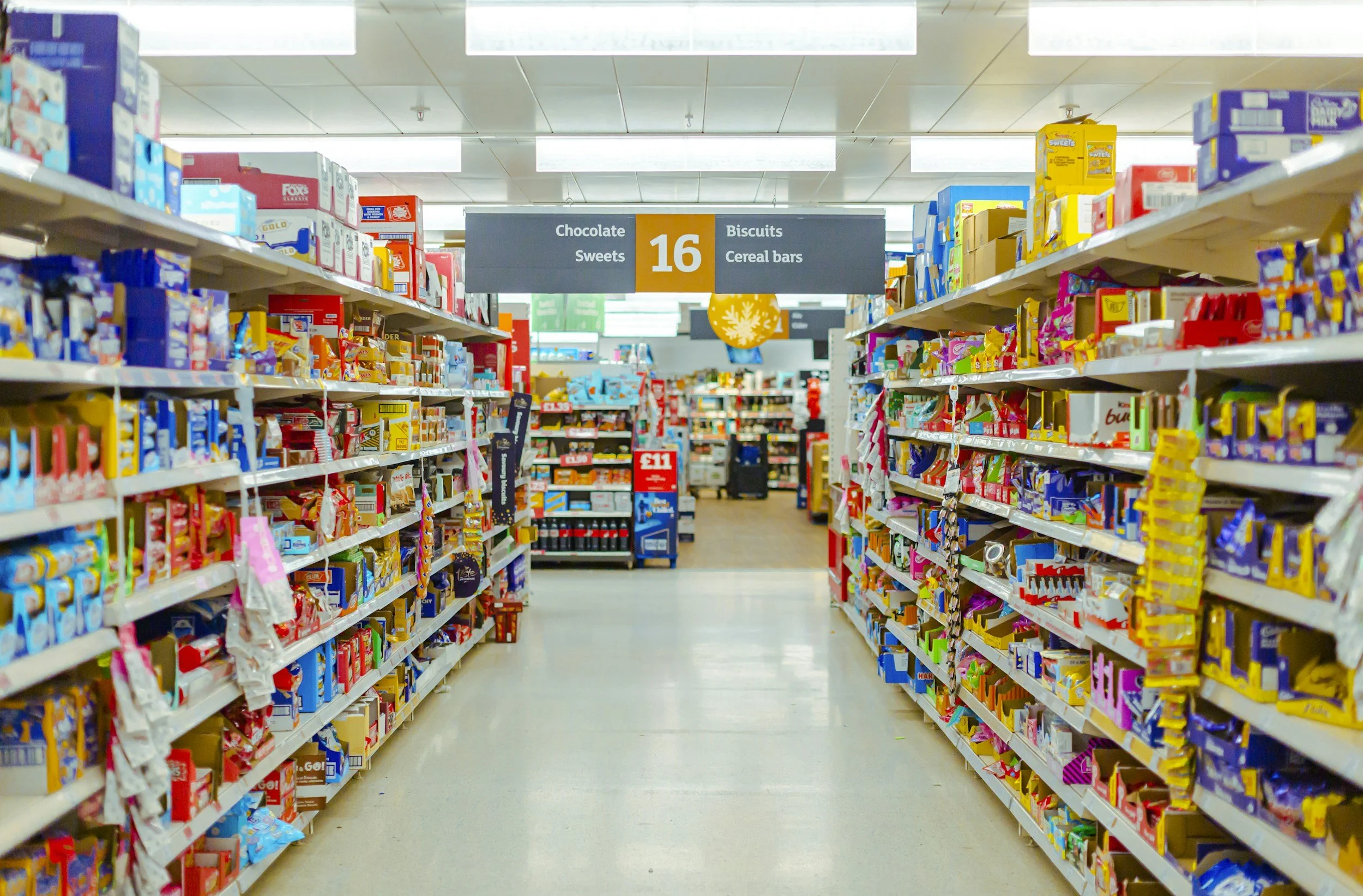 Aisle in a grocery store with shelves stocked with chocolate, sweets, biscuits, cereal bars, and other snacks, and a sign overhead indicating aisle 16.