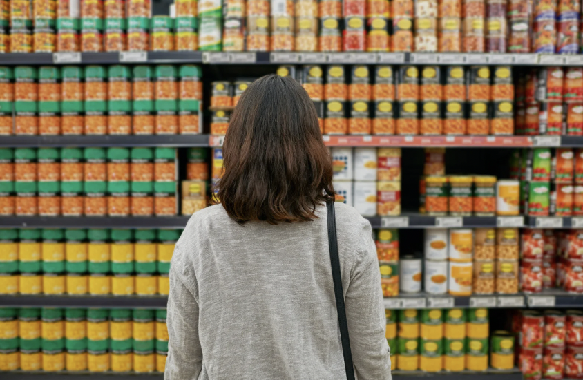 A woman with shoulder-length brown hair wearing a gray sweater and carrying a black shoulder bag, standing in front of grocery store shelves stocked with canned soup and other canned goods.