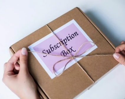 A person holding a brown subscription box wrapped with a string, labeled "Subscription Box" on a purple and white paper.