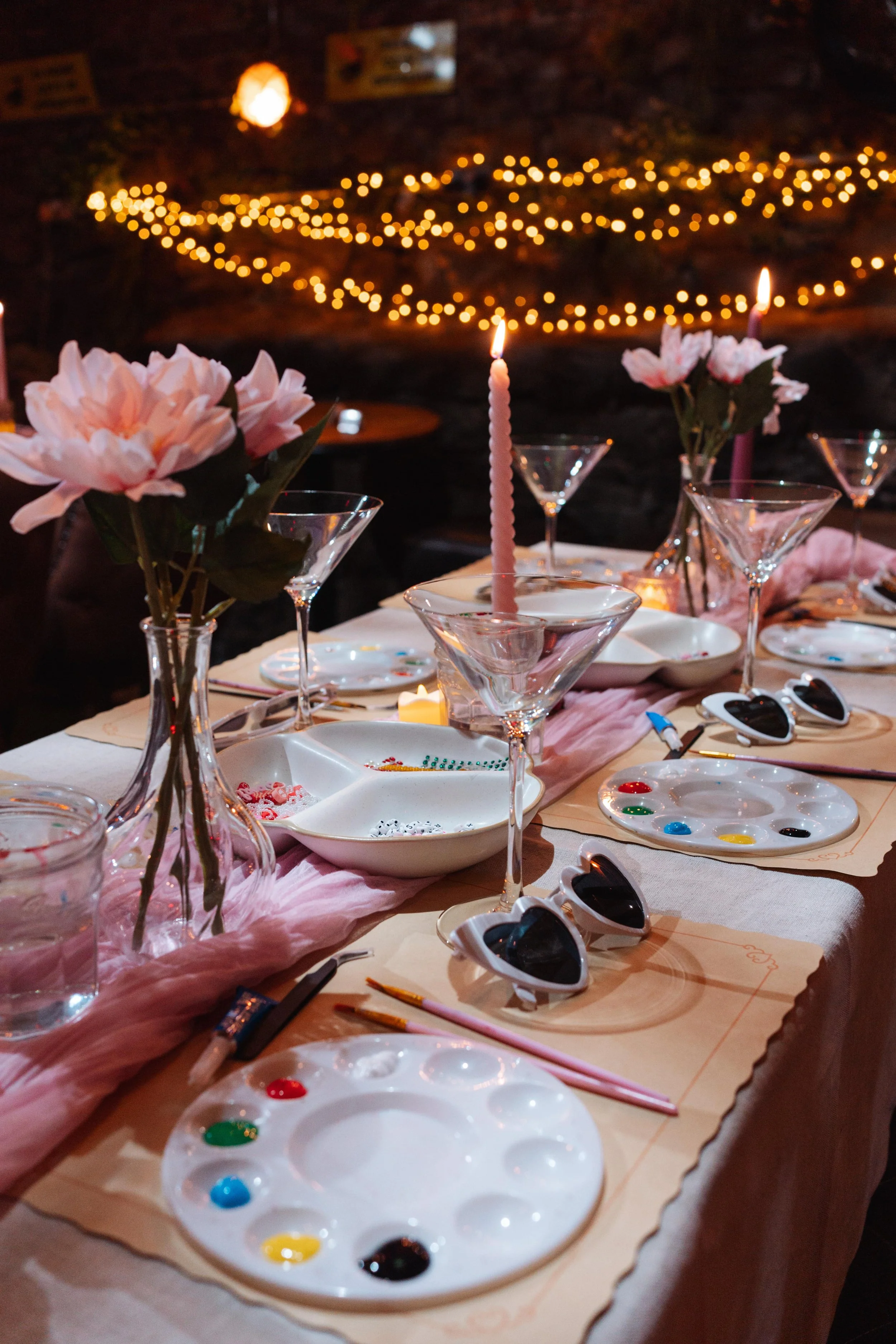 Decorated dinner table with pink flowers, candles, glasses, and paint palettes set for a painting party or event, with fairy lights in the background.
