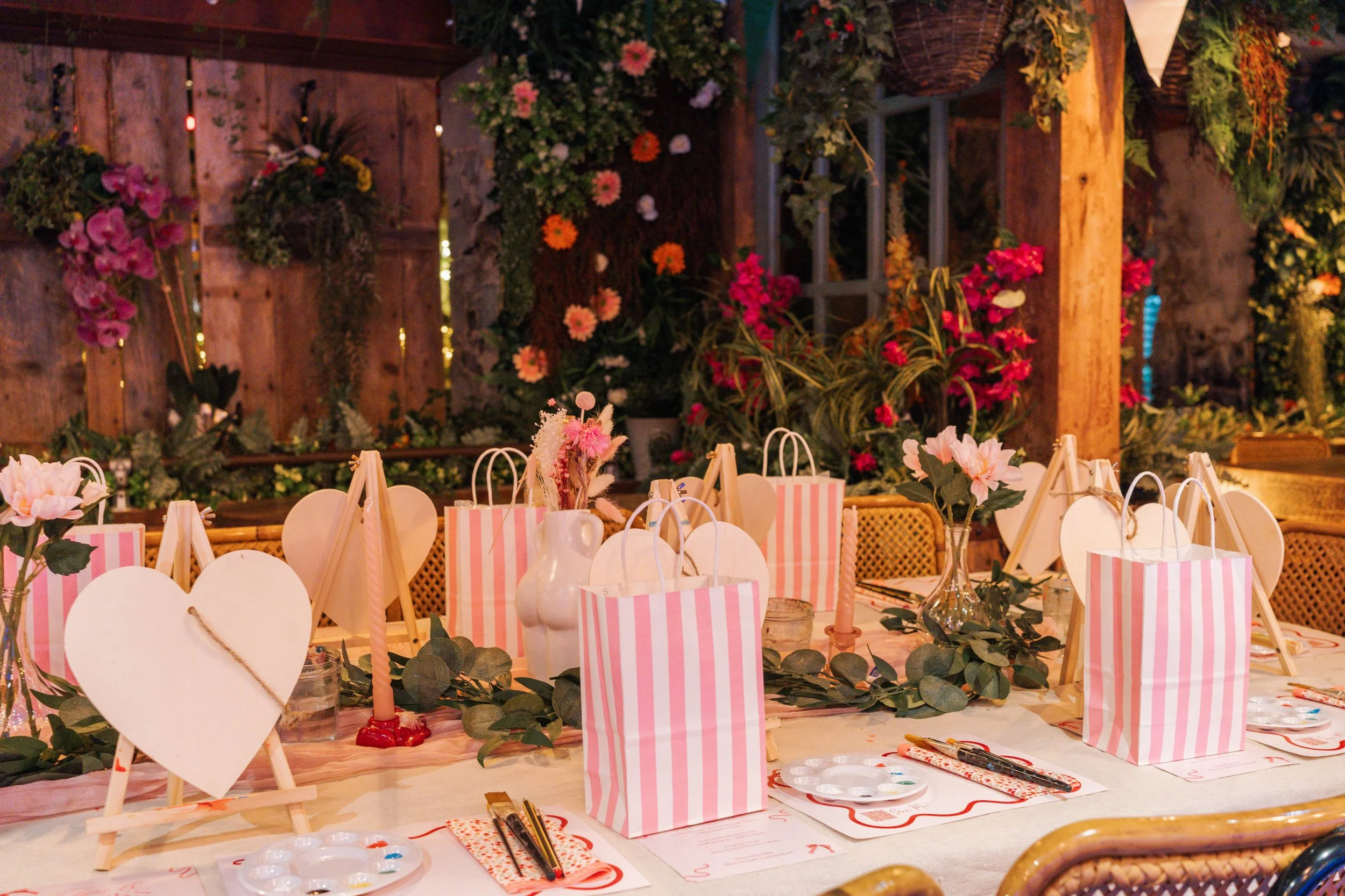 Decorated table with pink and white striped gift bags, candles, flowers, and greenery, set in a rustic, flower-filled setting for a celebration or gathering.