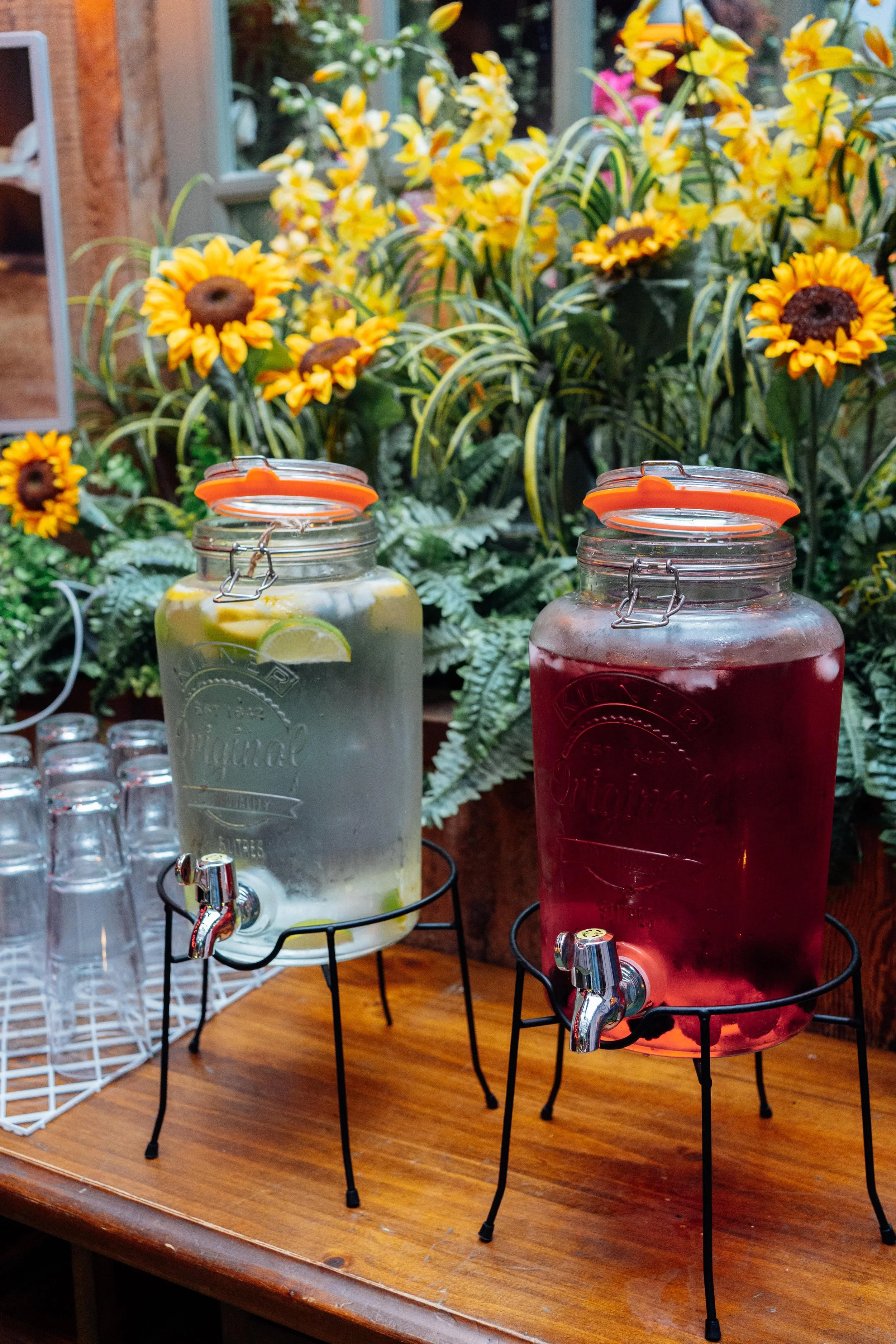 Two glass dispensers filled with lemonade and berry-infused water, placed on a wooden table with a background of yellow and orange flowers.