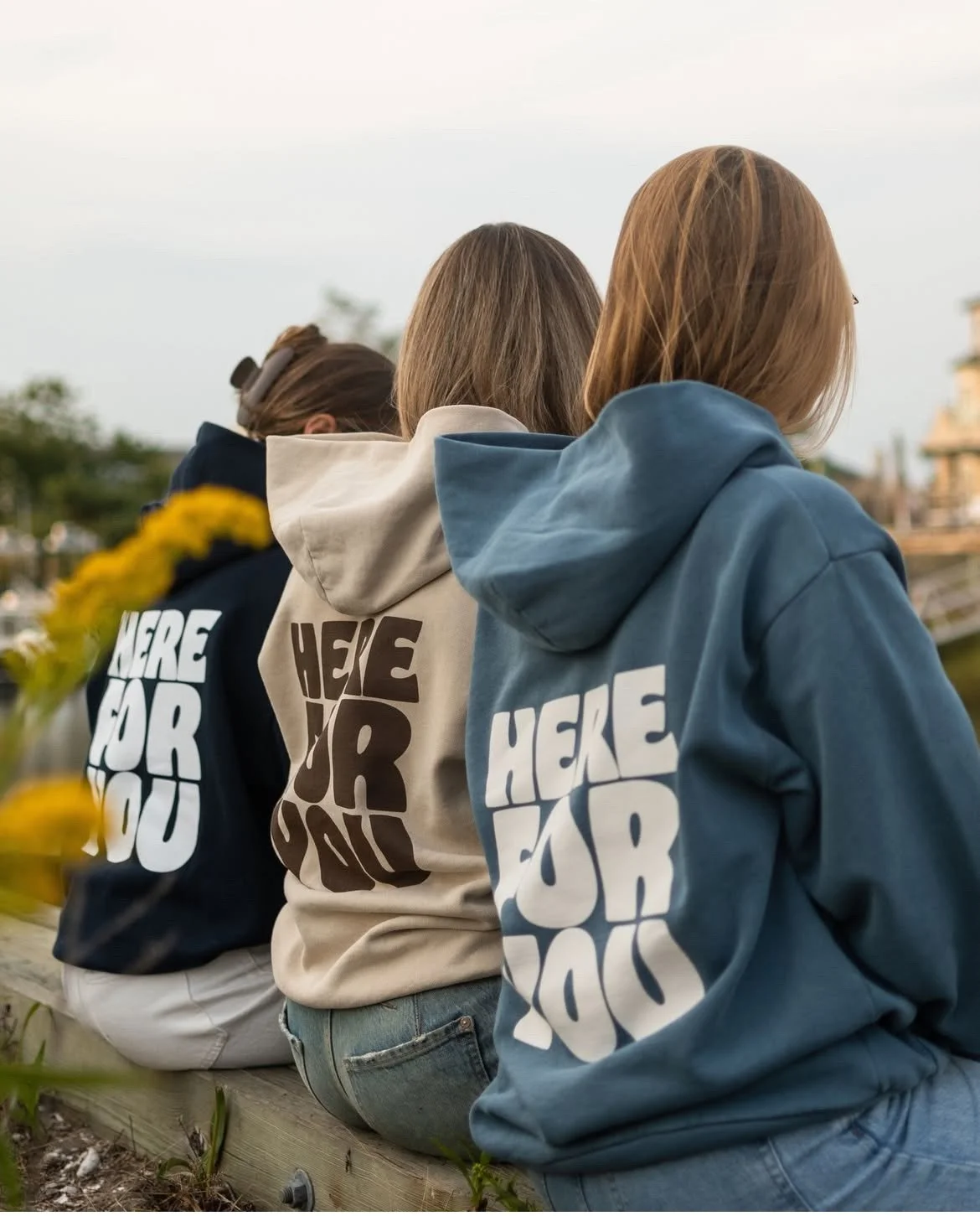 Three women sitting outdoors wearing hoodies with bold text on the back reading 'HERE FOR YOU'.