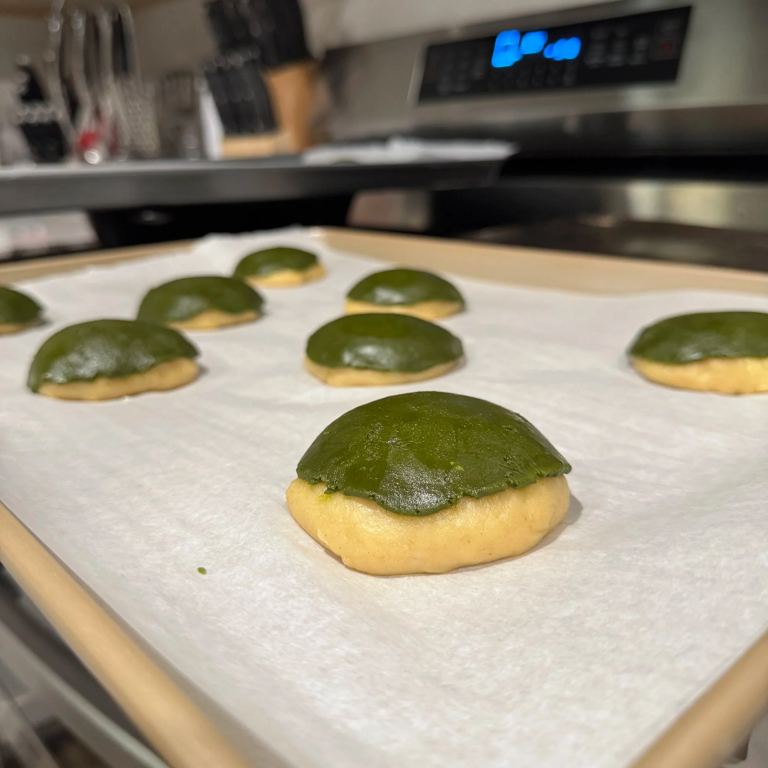 Mexican sweet bread with matcha streusel rising on a baking sheet lined with parchment paper in a kitchen.