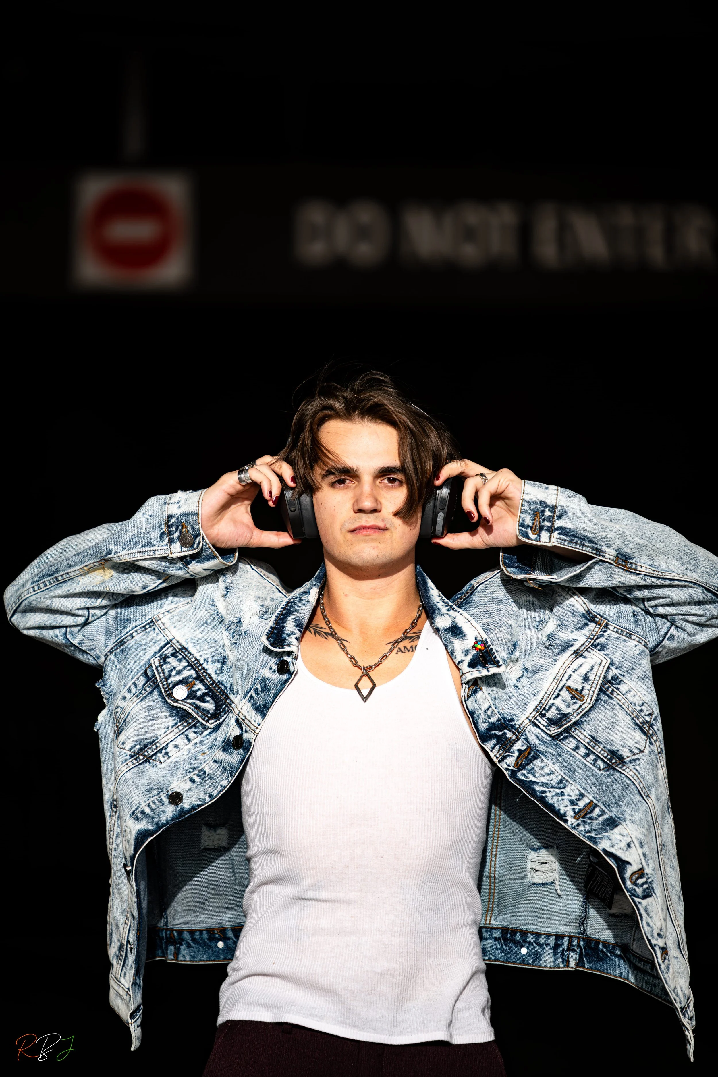 A person with shoulder-length dark hair wearing a light denim jacket over a white tank top, listening to headphones with a serious expression against a black background with a blurred 'Do Not Enter' sign.