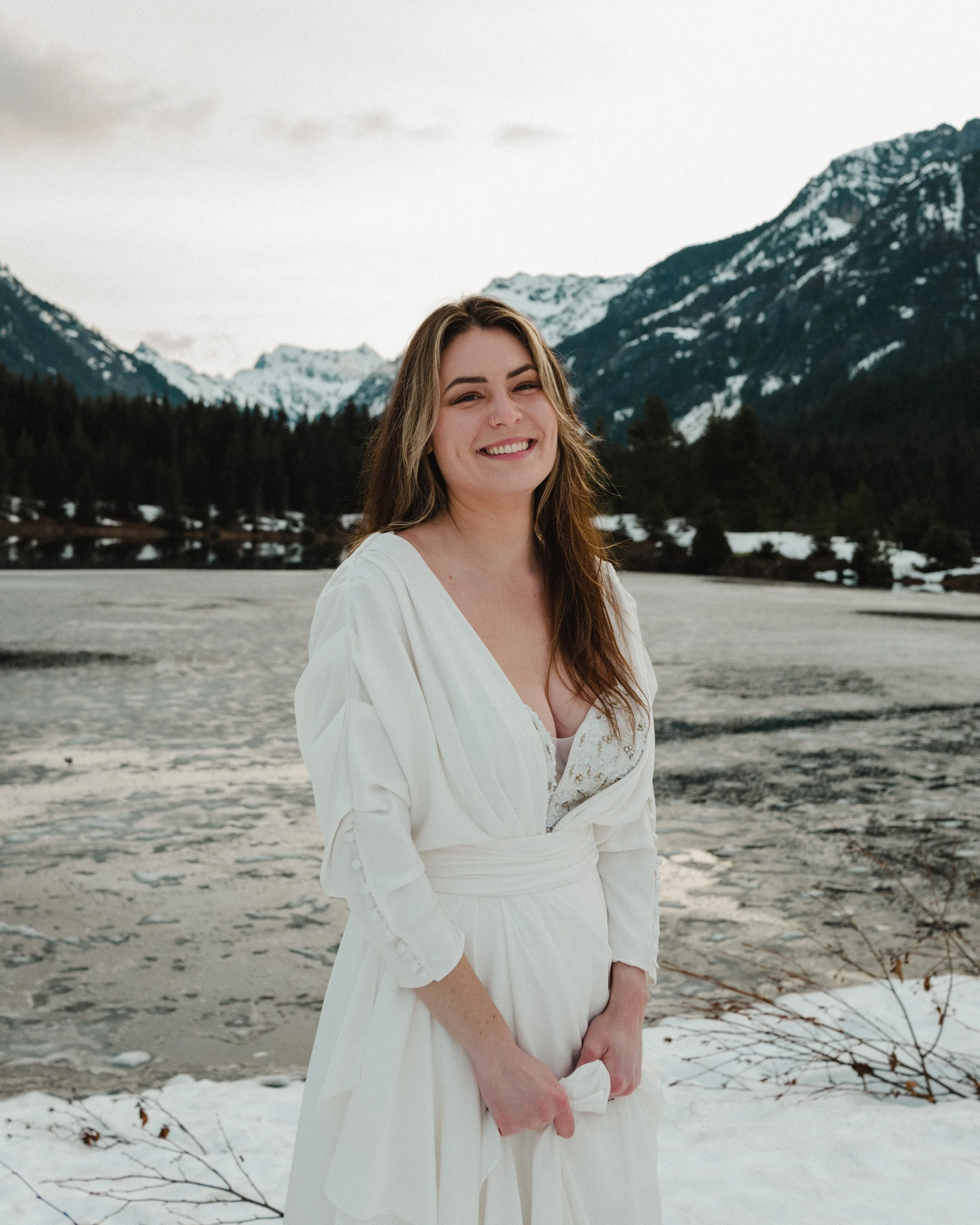 A woman in a white dress standing outdoors on snow with mountains in the background, smiling.