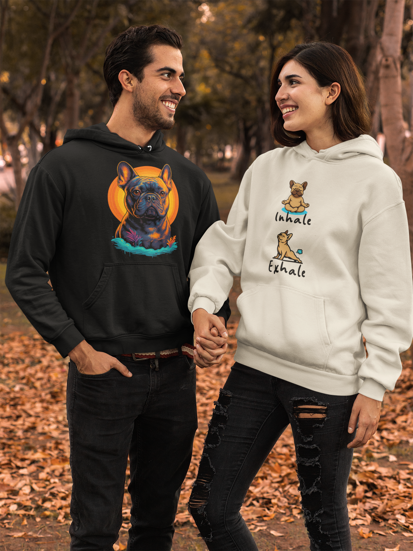 A smiling man and woman holding hands in an outdoor park during autumn. The man wears a black hoodie with a colorful French Bulldog graphic, and the woman wears a white hoodie with a dog illustration and the words "Inhale" and "Exhale."