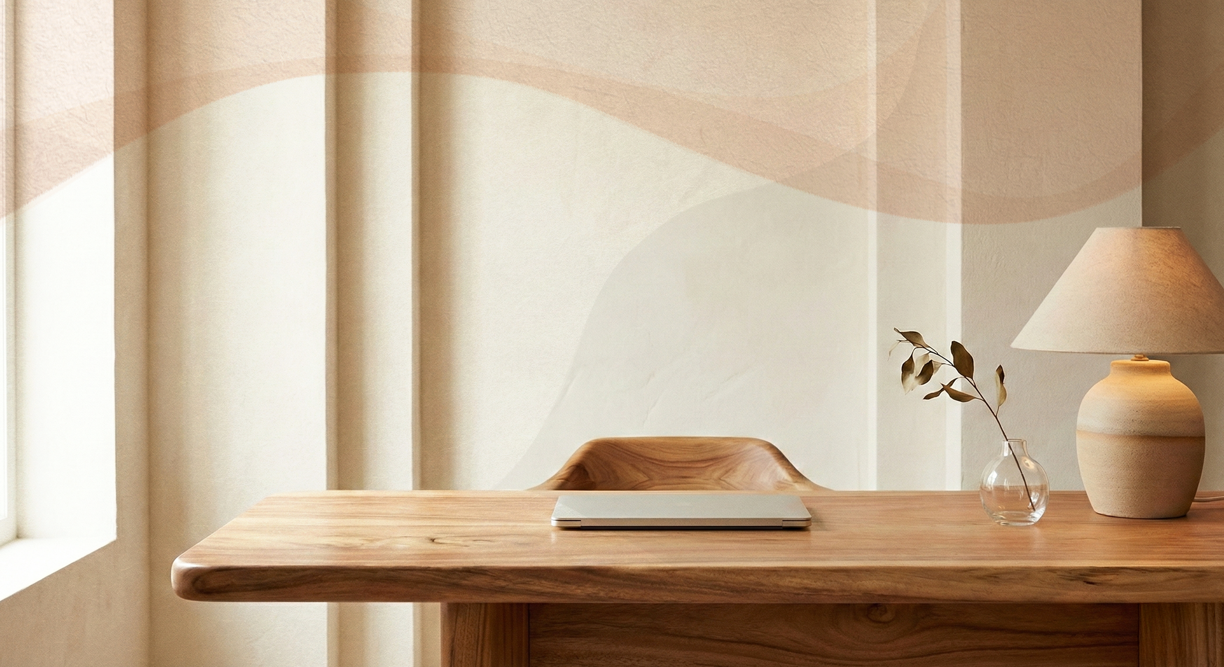 A minimalist wooden desk with a closed laptop, a small glass vase with a branch of leaves, and a large beige table lamp in a room with beige curtains and textured wall.