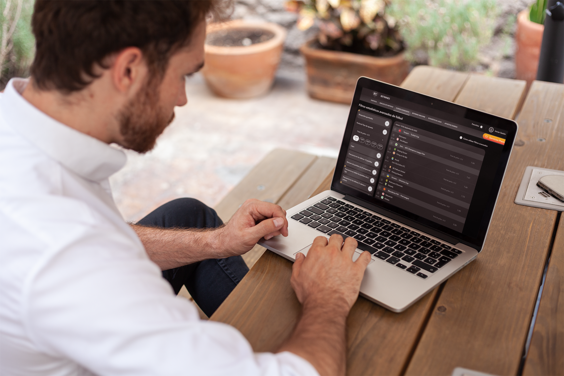 A man with brown hair and beard sitting at a wooden table using a silver MacBook laptop displaying a dark-themed sports analytics website with statistics and filters, outdoors with potted plants in the background.
