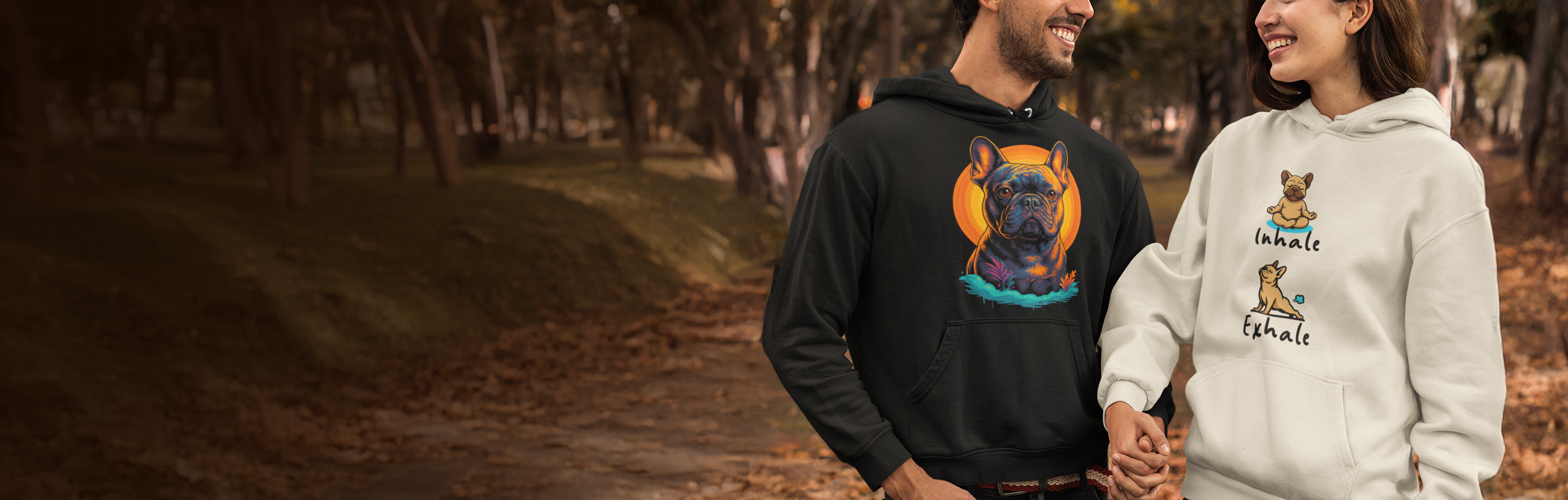 A couple holds hands and smiles at each other in an outdoor park with trees during autumn. They wear hoodies with dog illustrations and words, the man's black hoodie features a French Bulldog, and the woman's cream hoodie has cartoon dogs with the words "Inhale" and "Exhale."