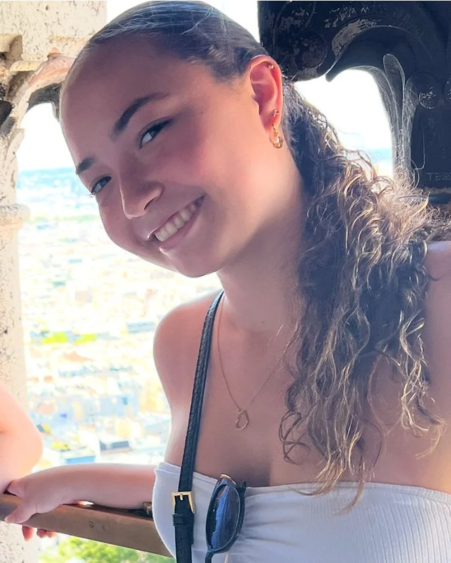 A young woman with curly hair, earrings, and a necklace, smiling for a photo outside with a city view in the background.