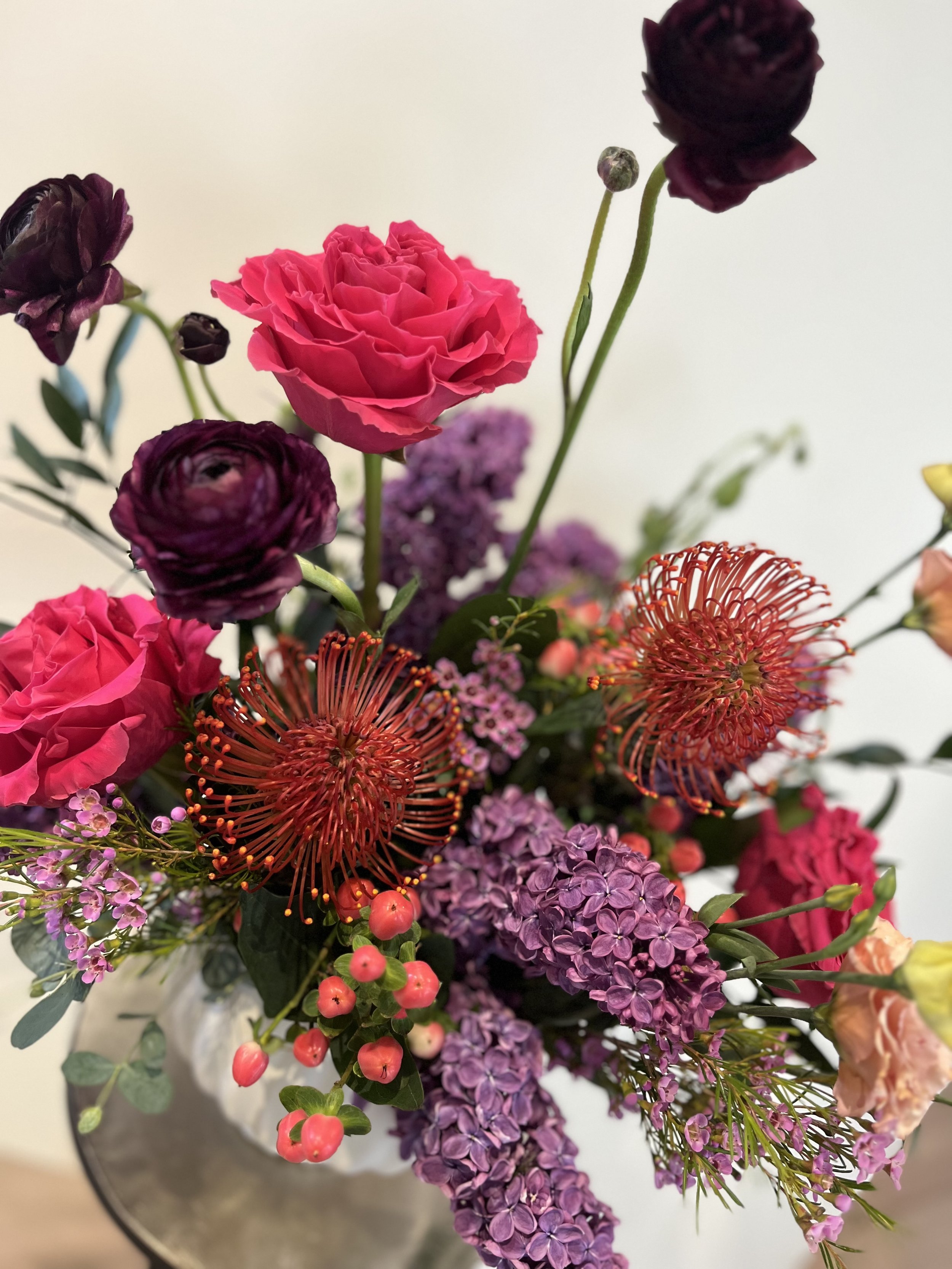 A vibrant floral arrangement featuring pink roses, purple ranunculus, red pincushion protea, lilac flowers, hypericum berries, and assorted greenery.