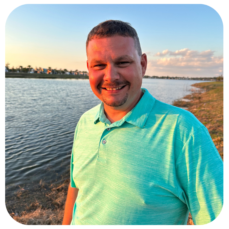 A man smiling outdoors near a body of water during sunset, wearing a light green polo shirt.