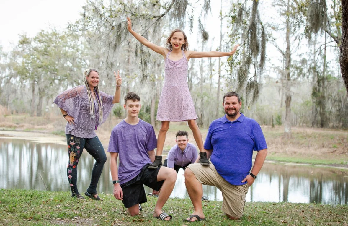 A family of five posing outdoors near a pond, with one girl balancing on their shoulders, all smiling and making peace signs.