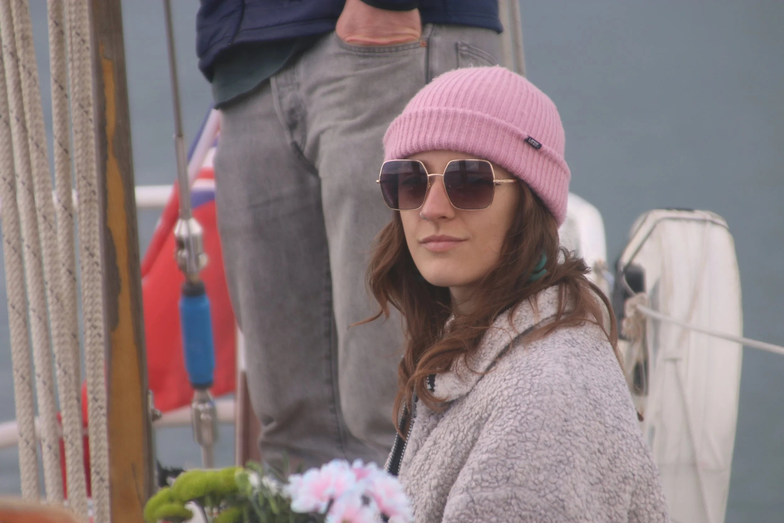 A young woman with long brown hair wearing sunglasses, a pink knit beanie, and a fleece jacket, is standing outdoors near a boat, possibly on a dock, with a woman in the background. The background includes ropes and boating equipment.