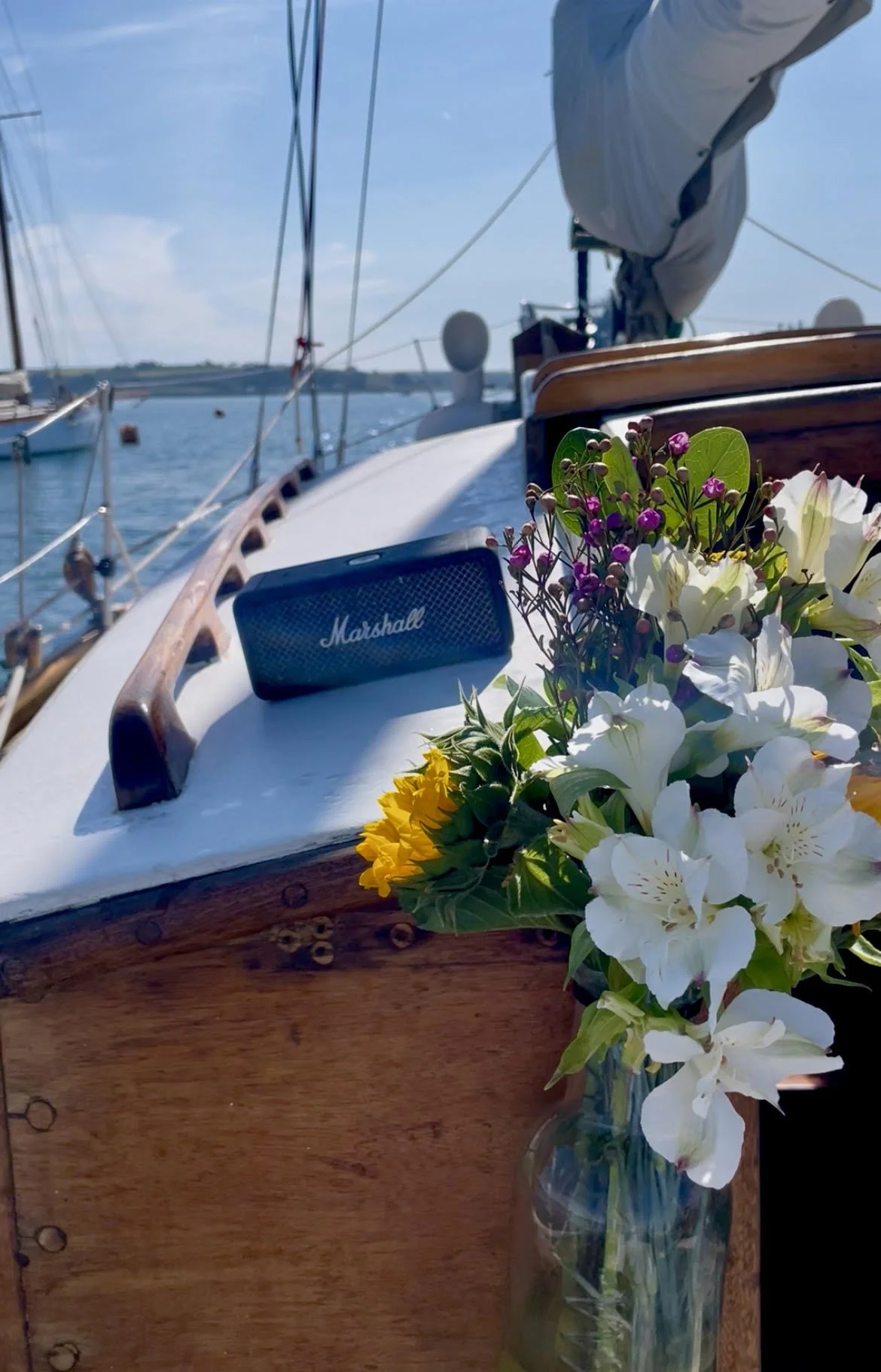 A bouquet of white, purple, and yellow flowers in a glass vase placed on the wooden edge of a boat. A portable black Marshall speaker is also on the boat's surface. The background shows sailboat masts and calm water under a partly cloudy sky.