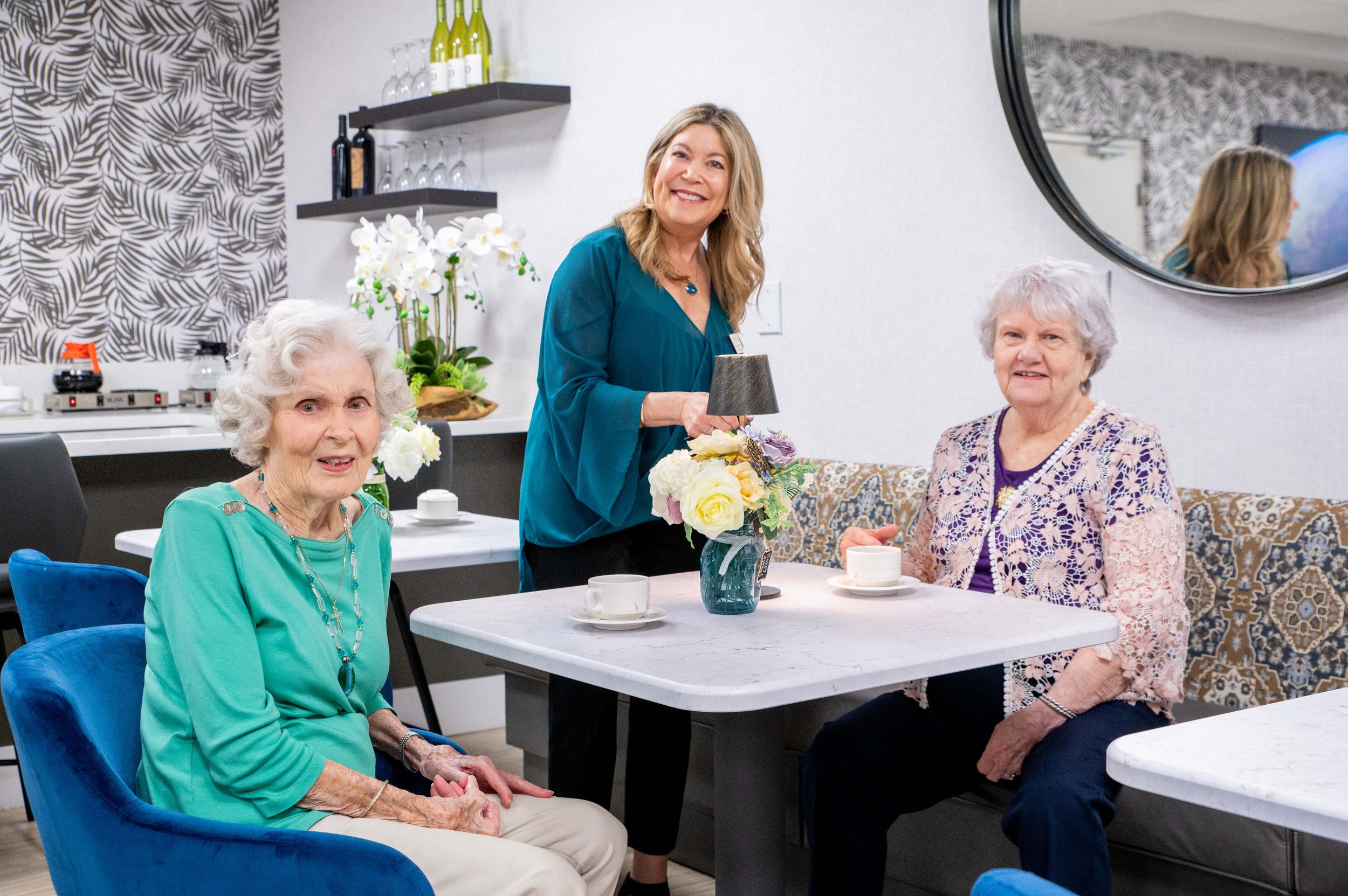 Two elderly women sitting at a table with a younger woman standing between them in a stylish cafe setting, with decorative wallpaper, a mirror, and a shelf of wine bottles in the background. There are flowers on the table, along with teacups and saucers.
