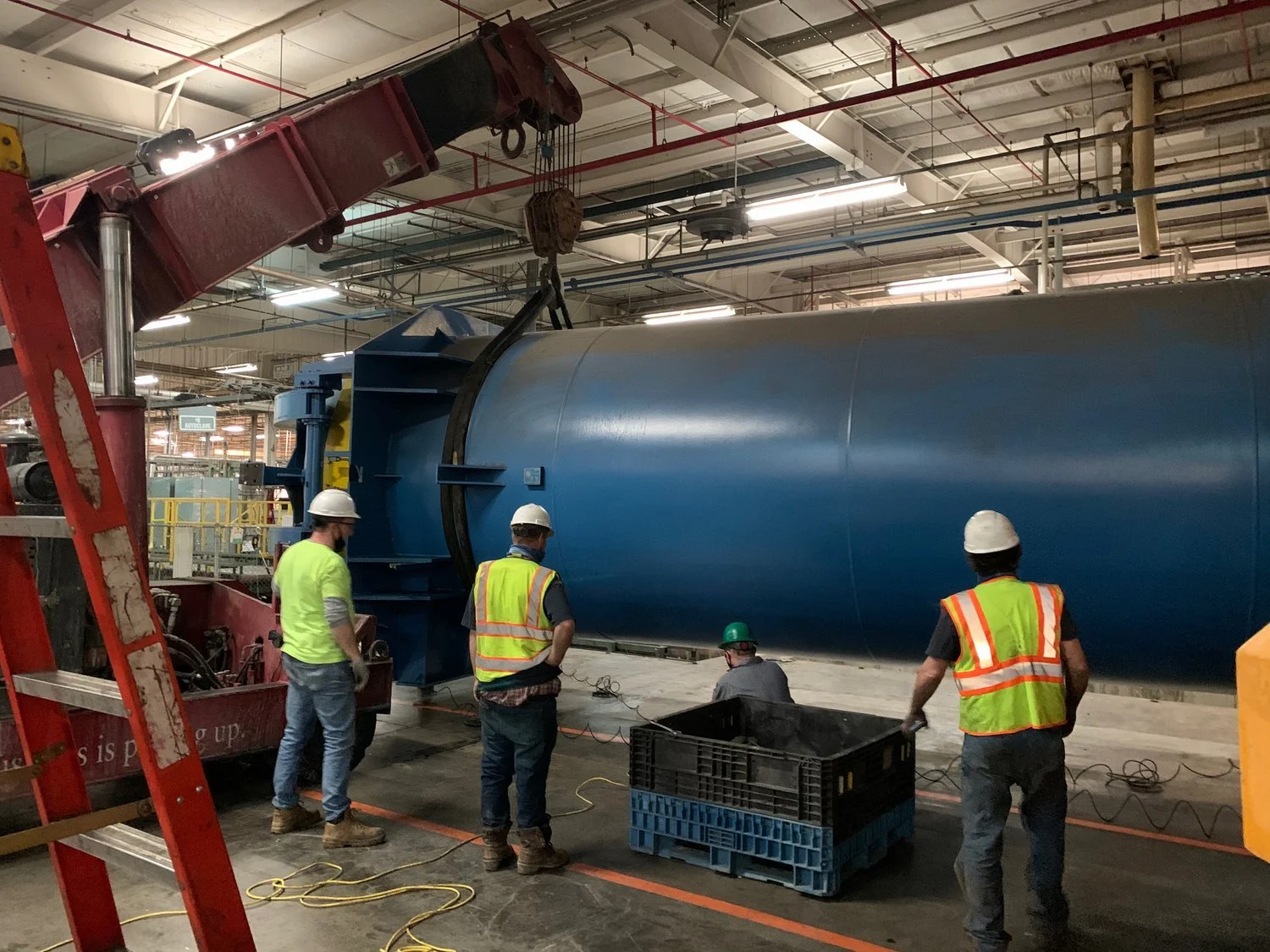 Workers in safety helmets and vests working on a large blue turbine or industrial equipment inside a factory or industrial facility, with a crane nearby.