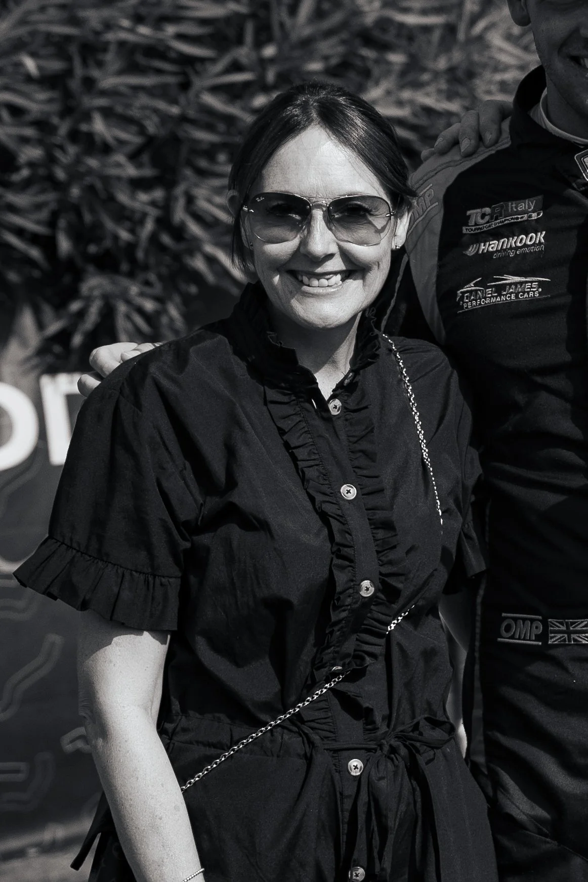 A smiling woman wearing sunglasses and a black button-up shirt with ruffles, standing outdoors.
