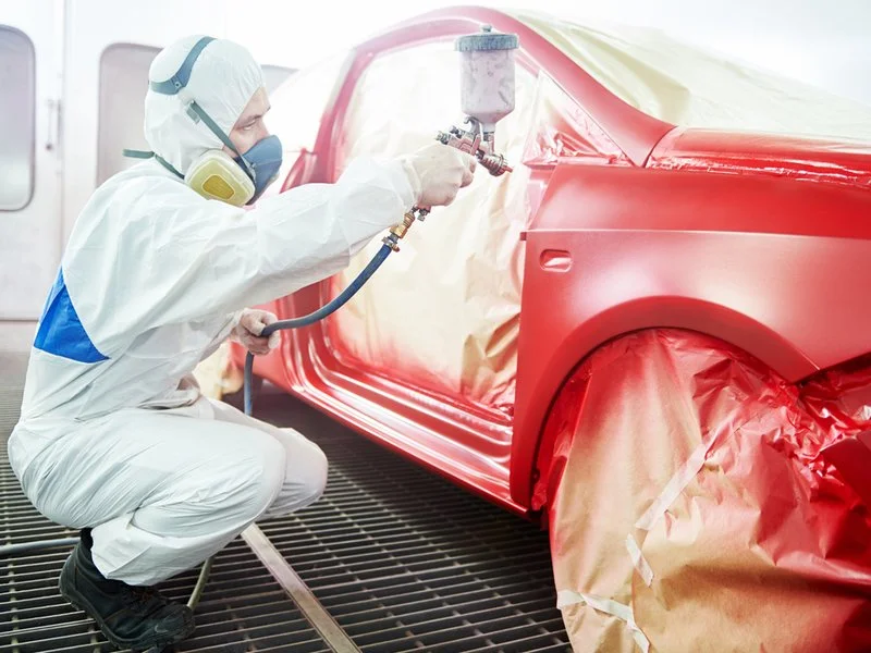 A worker in protective gear spray painting a red car in a manufacturing or repair facility.