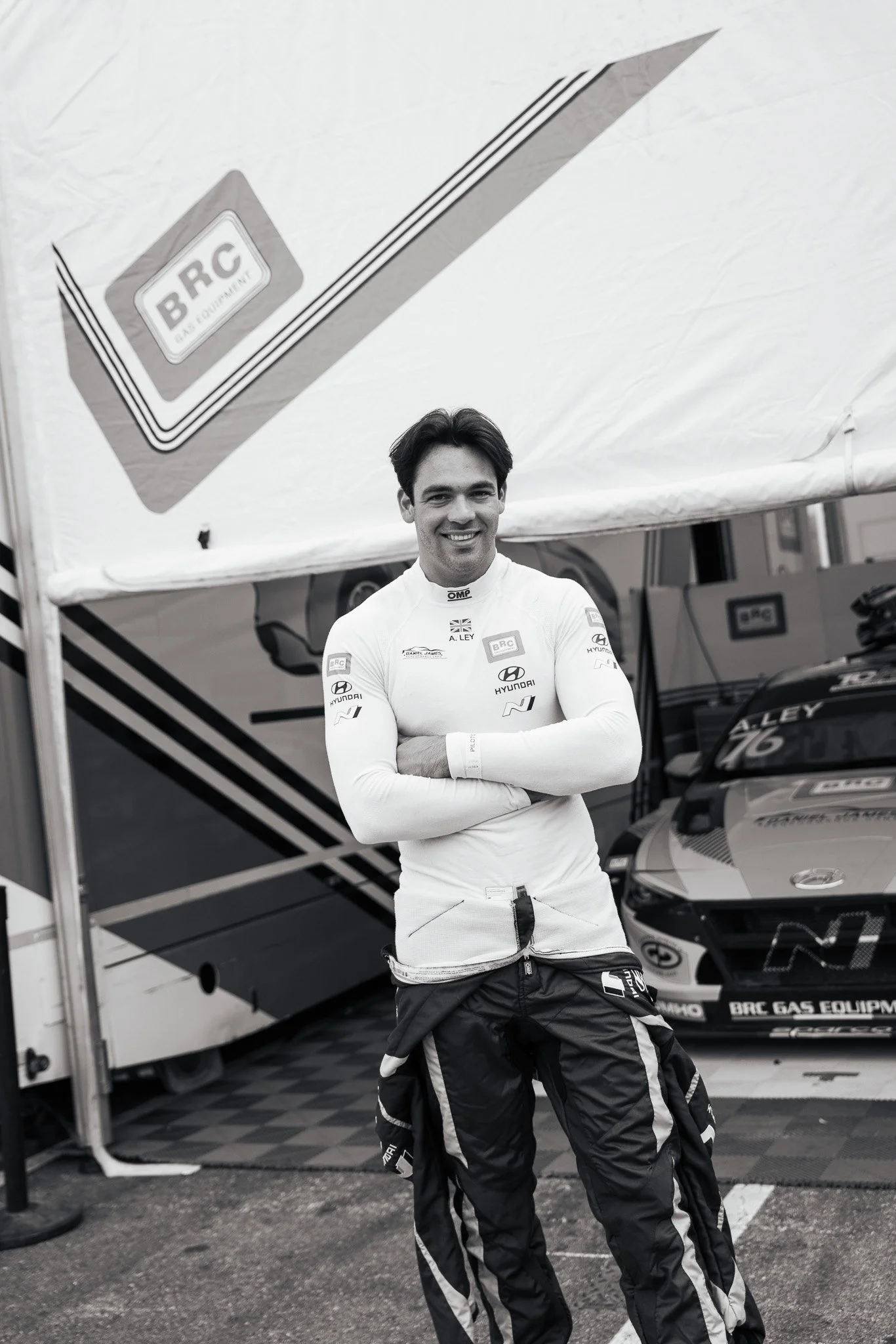A male race car driver standing in front of a race car under a canopy in a racing paddock area.