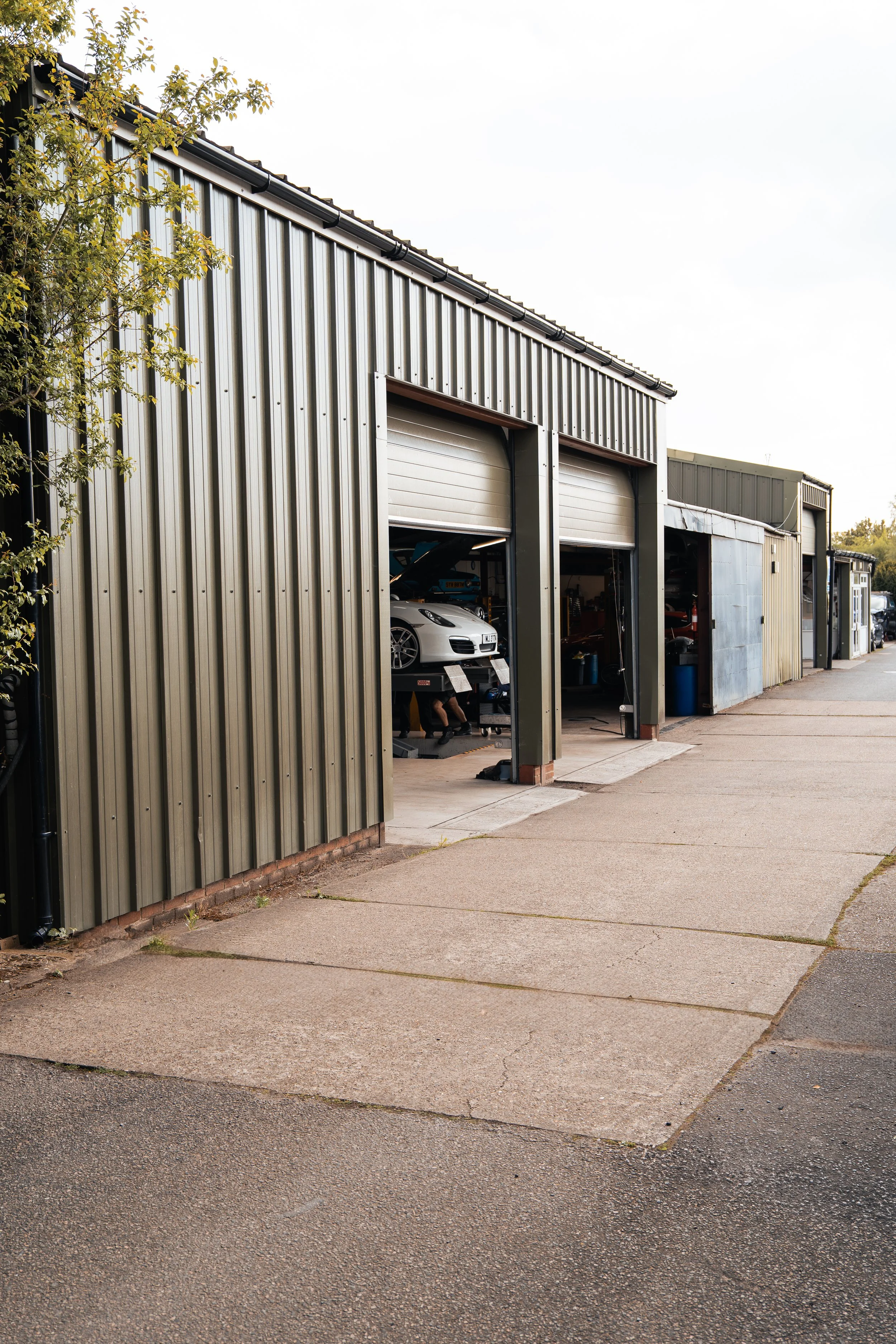 An automotive repair shop with metallic exterior walls and partially open garage doors, revealing a white sports car on a lift inside.