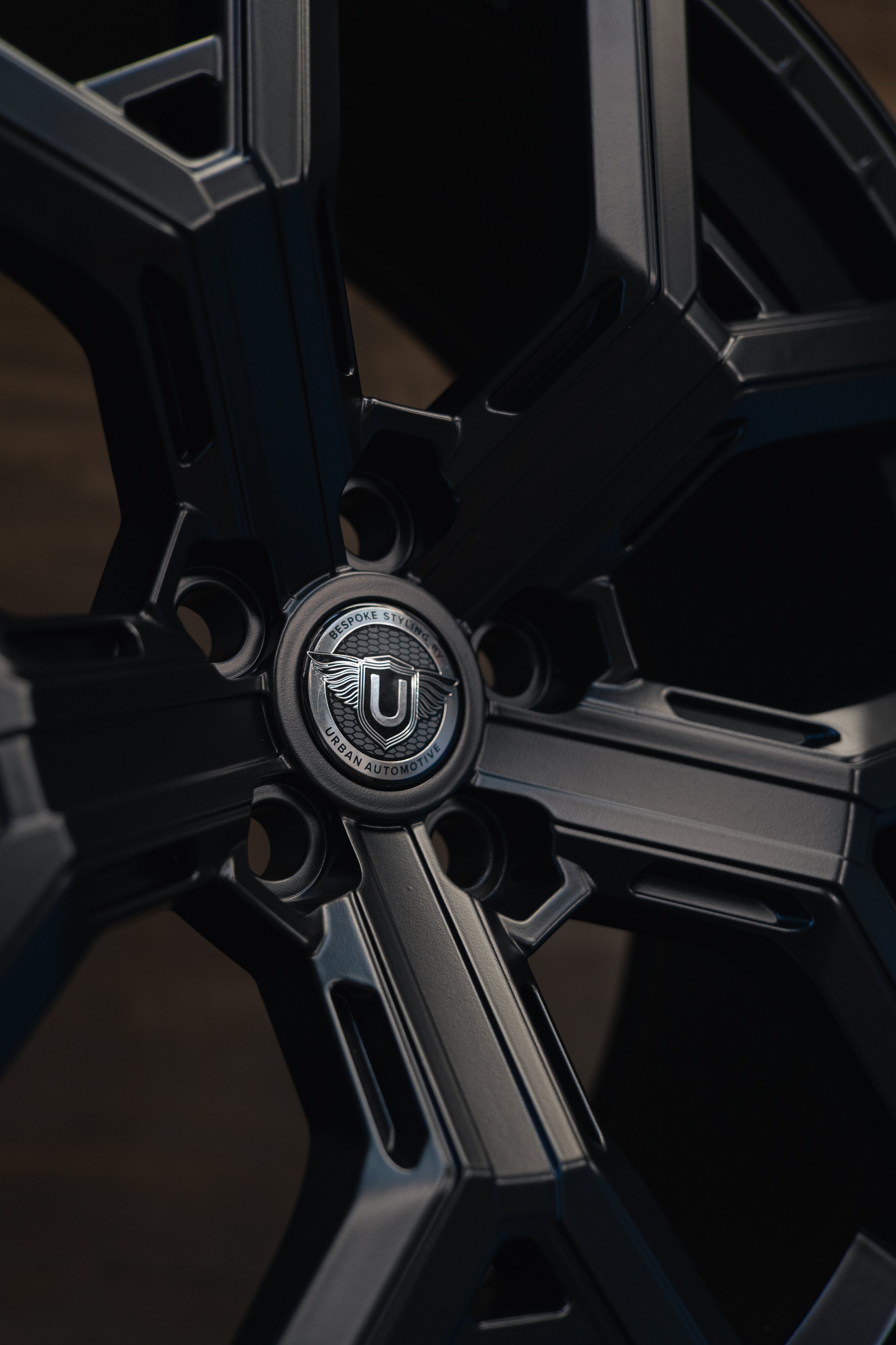 A close-up of a black, multi-spoke car wheel with a central logo reading 'U' and 'Urban Automotive,' featuring wings, on a dark background.
