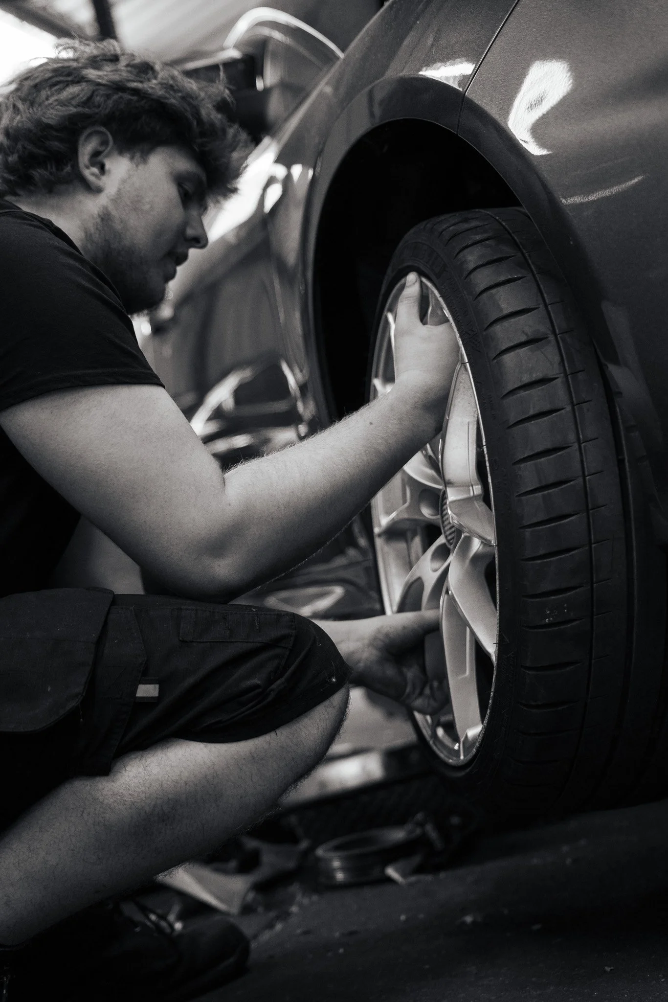 A young man with curly hair and a beard changing a flat tire on a car in a garage.