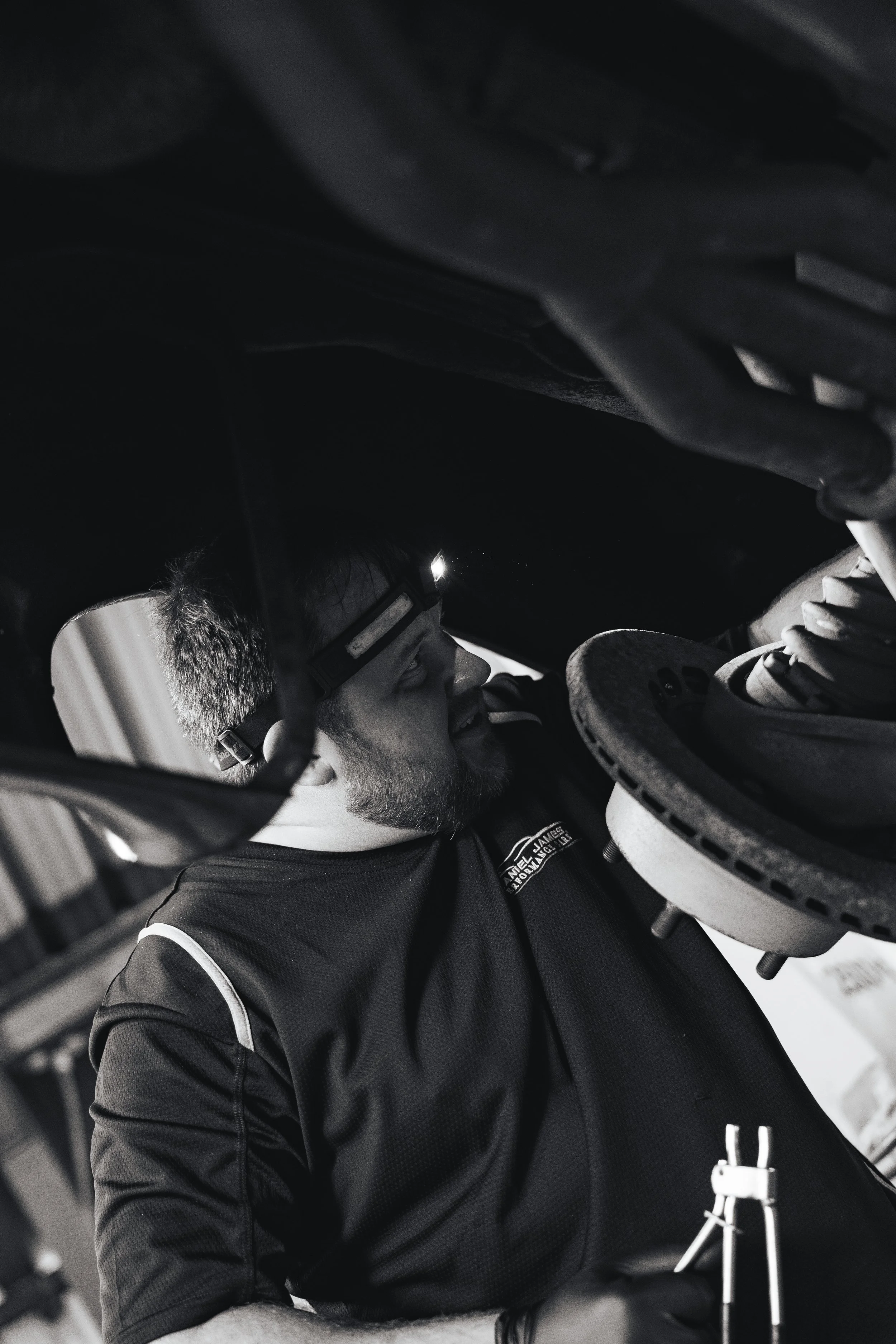 A man with a beard and glasses, wearing a headlamp, working under a vehicle in a garage.
