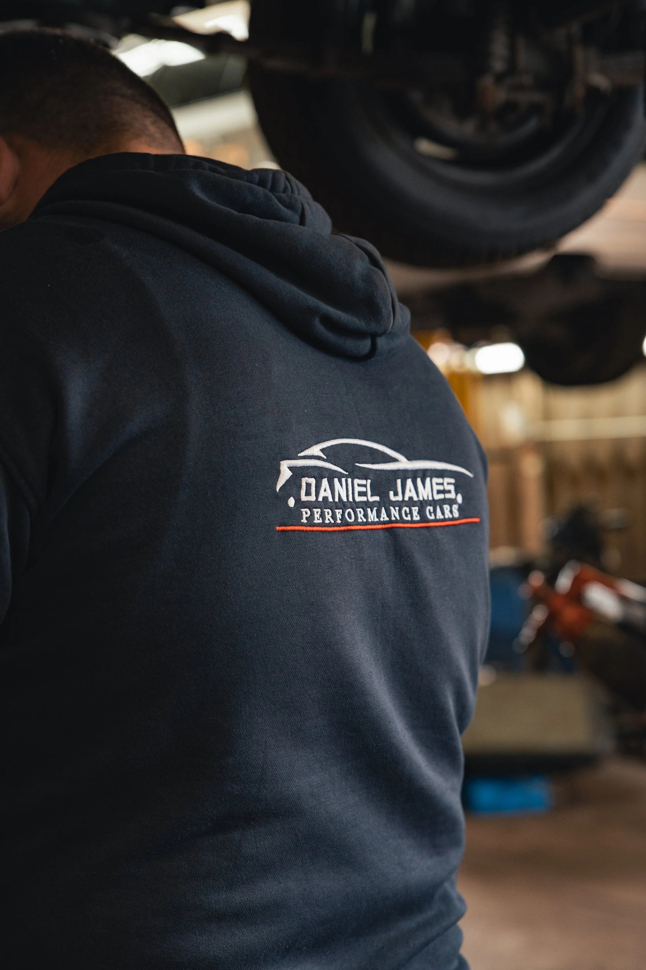 A person wearing a dark hoodie with a logo that reads "Daniel James Performance Cars" on the back, in an automotive workshop with a car lift overhead.