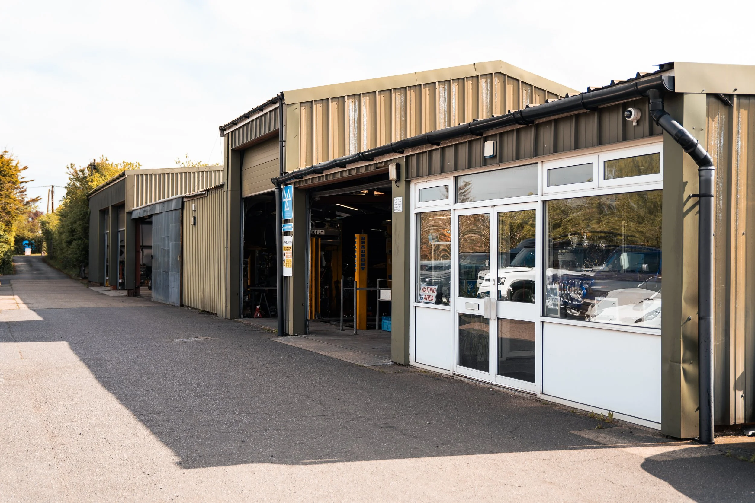 Auto repair shop with open garage bays and a glass door, cars inside and outside, and trees in the background.