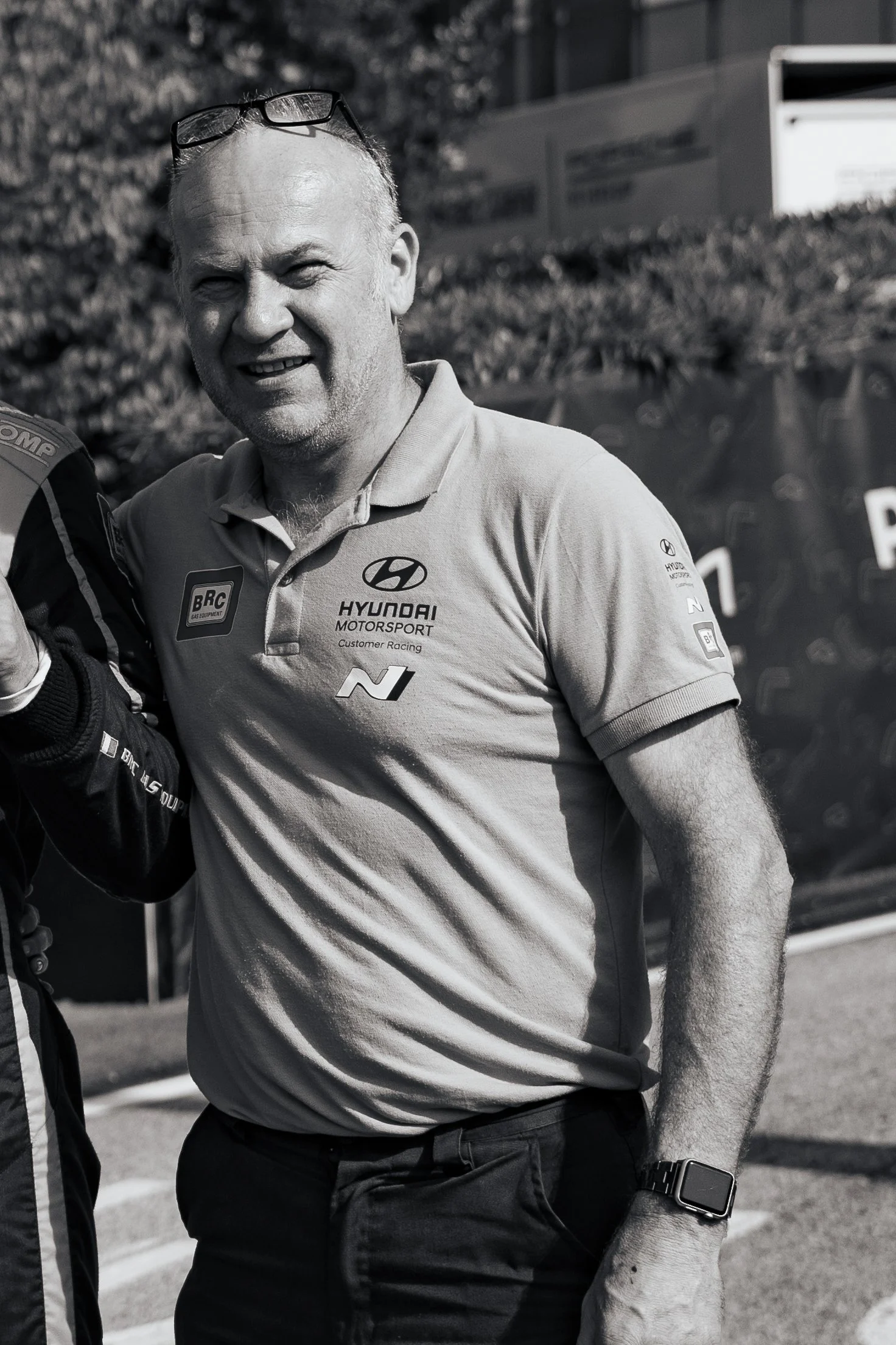 A smiling man wearing a Hyundai Motorsport polo shirt with various sponsor logos, sunglasses on his head, and a smartwatch on his wrist, standing outdoors at a racing event.