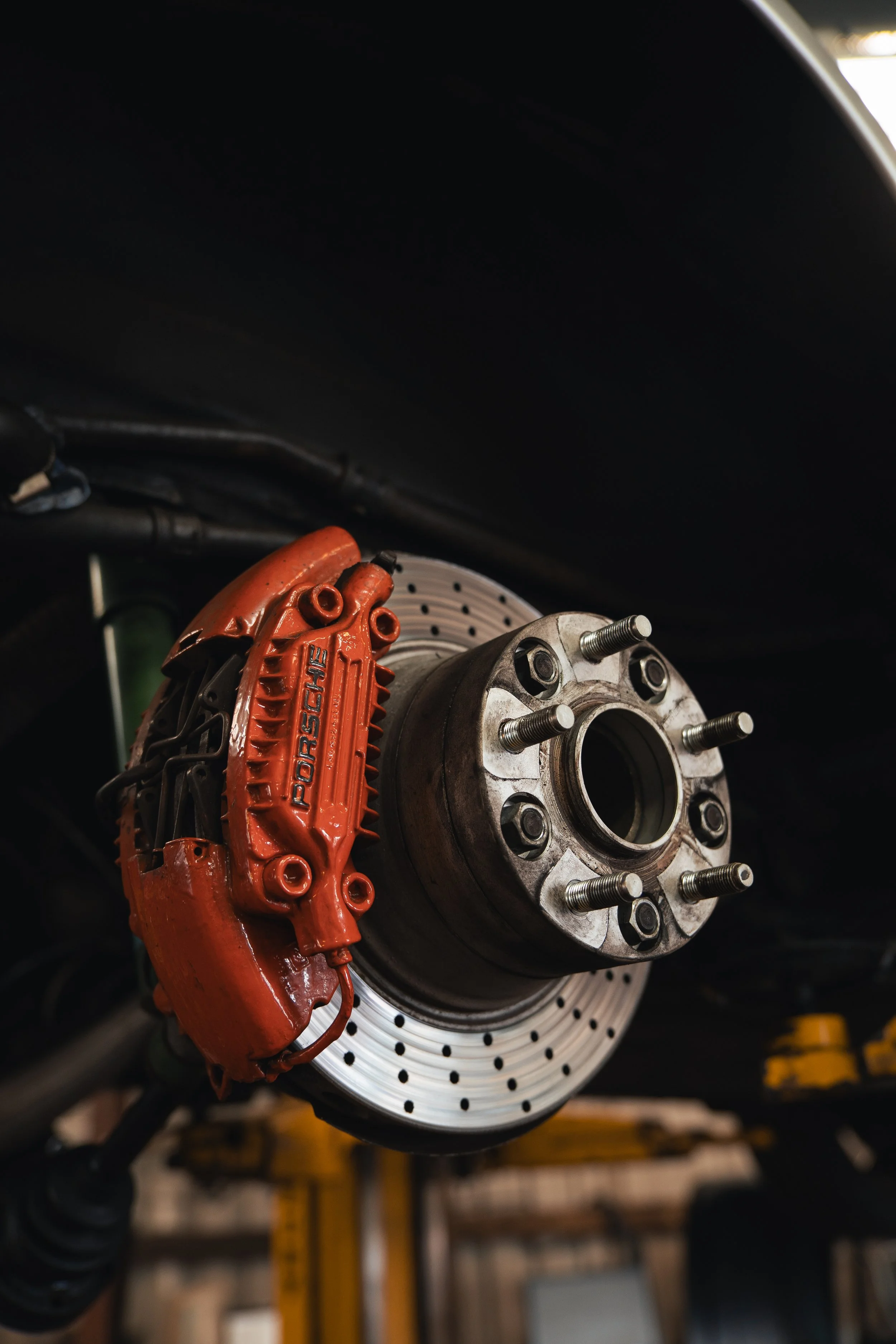 Close-up of a Porsche brake caliper and rotor on a car's wheel hub, with the car elevated on a hydraulic lift in a garage.