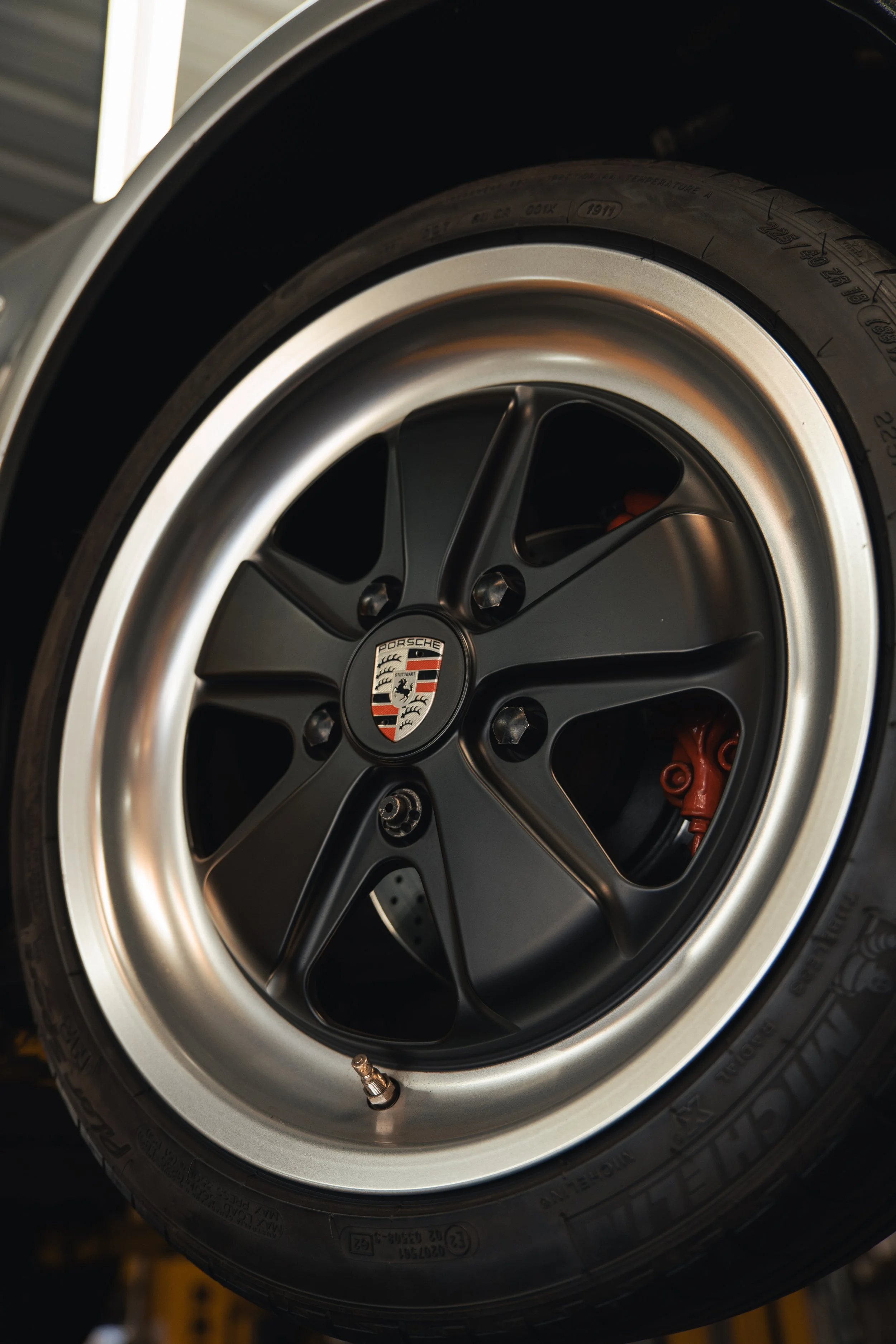 Close-up of a Porsche car wheel with a black rim, silver lip, and Porsche emblem in the center.