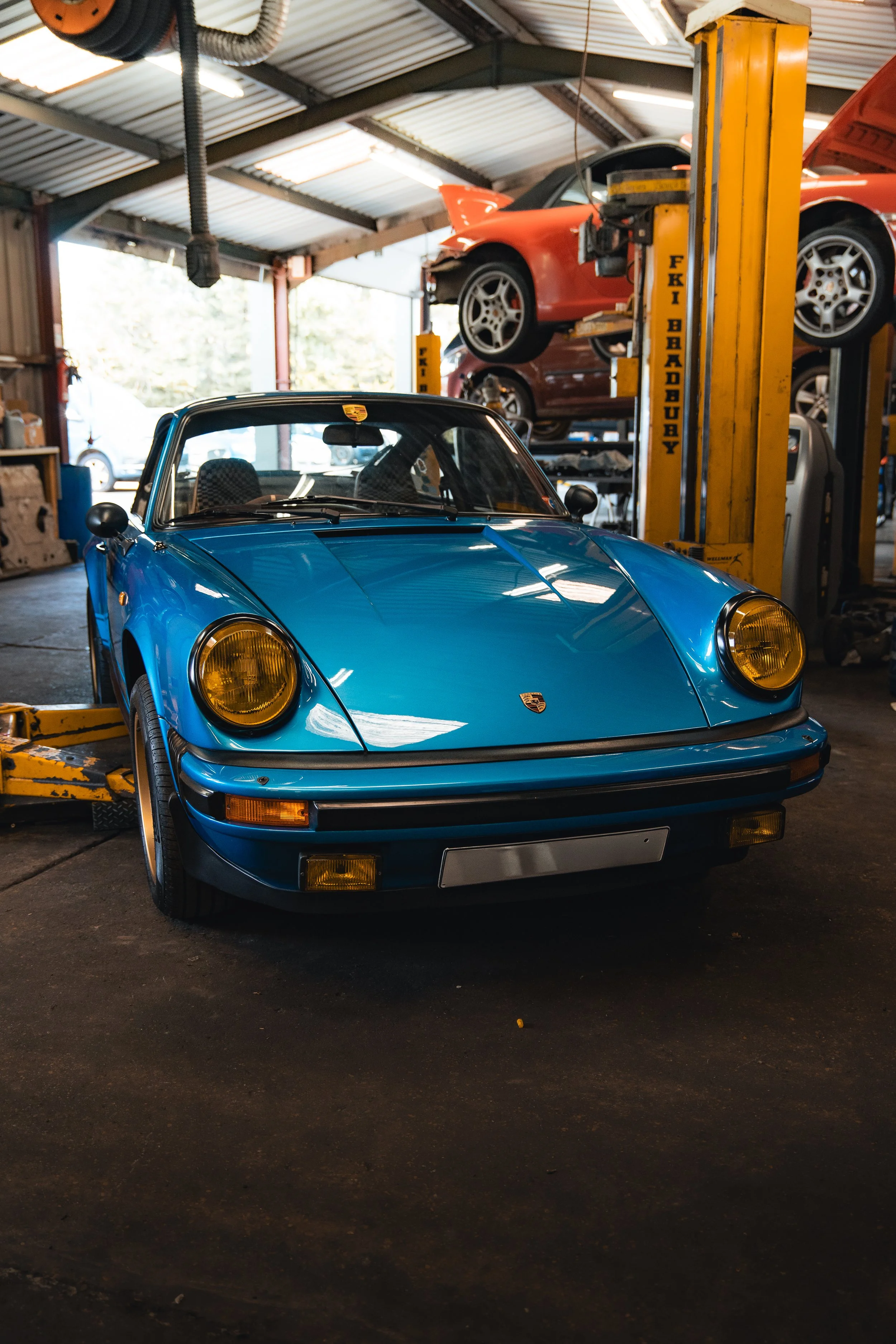 Blue classic Porsche sports car inside a garage or auto repair shop, with a yellow car lift and other cars in the background.