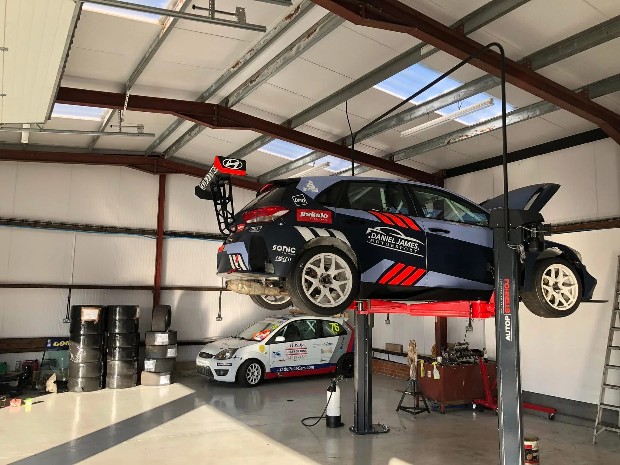 A race car is elevated on a hydraulic lift inside a garage, with another small race car parked nearby. The garage has tire stacks, a ladder, and tools, indicating a racing workshop.