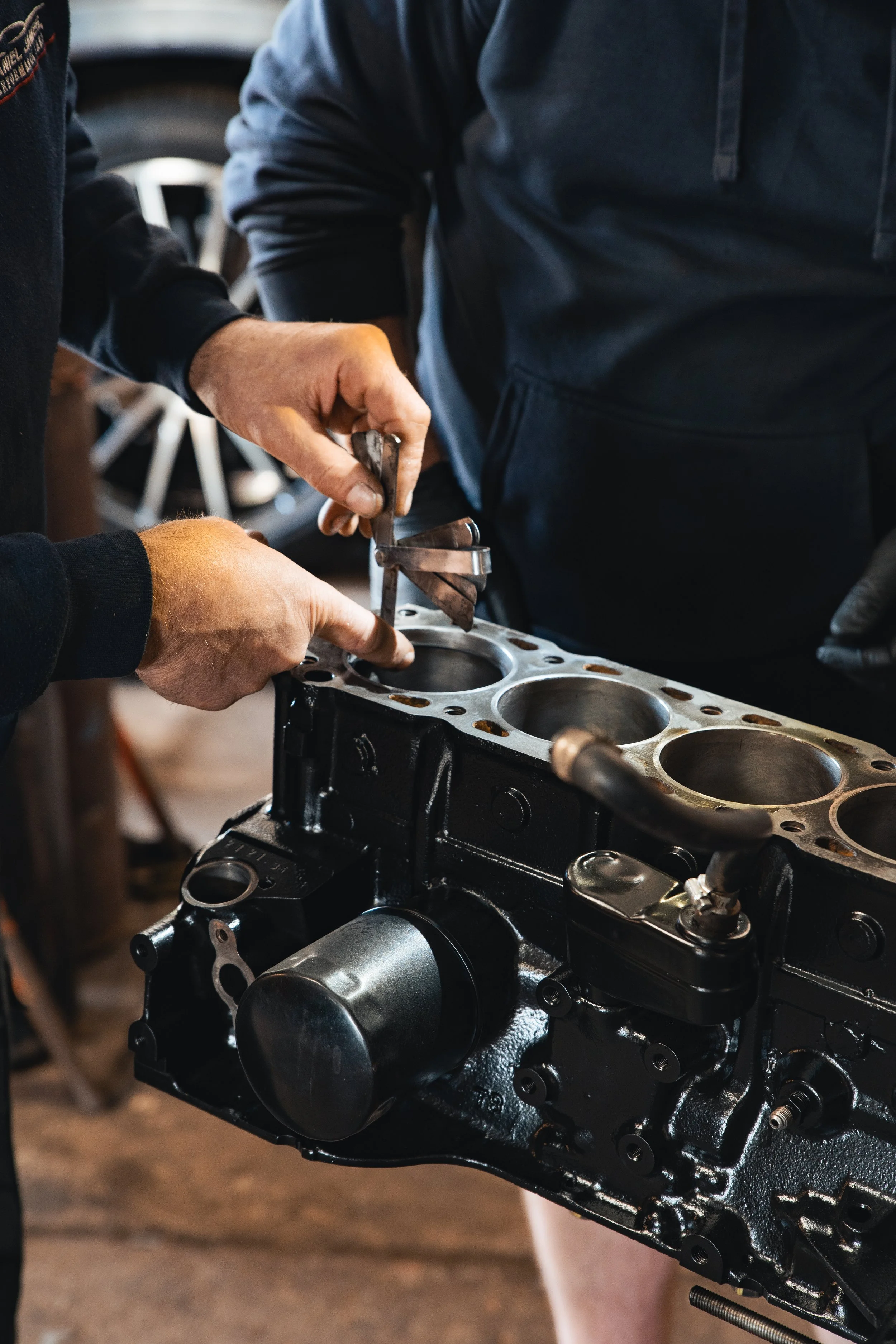 Two people working on an engine block, one using a tool to measure or adjust part of the engine in a workshop.