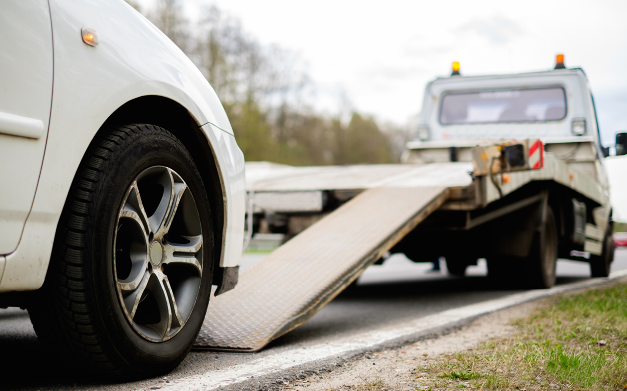 A white tow truck with a flatbed partly lowered on the side of a road, ready to tow a white vehicle.
