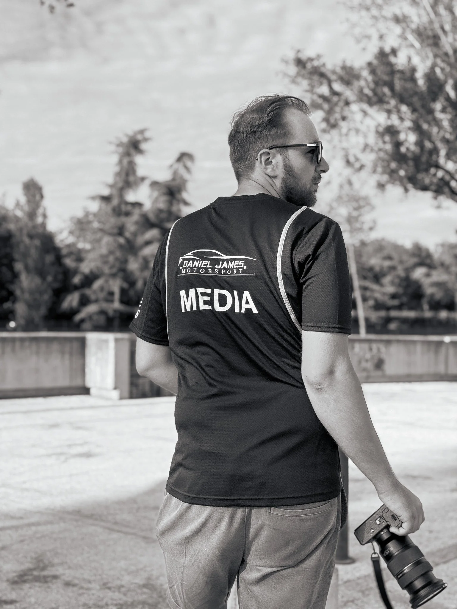 Photographer in black t-shirt with 'MEDIA' and 'DANIEL JAMES MOTORSPORT' logos, holding a camera with a large telephoto lens, standing outdoors with trees and a fence in background, black and white photo.