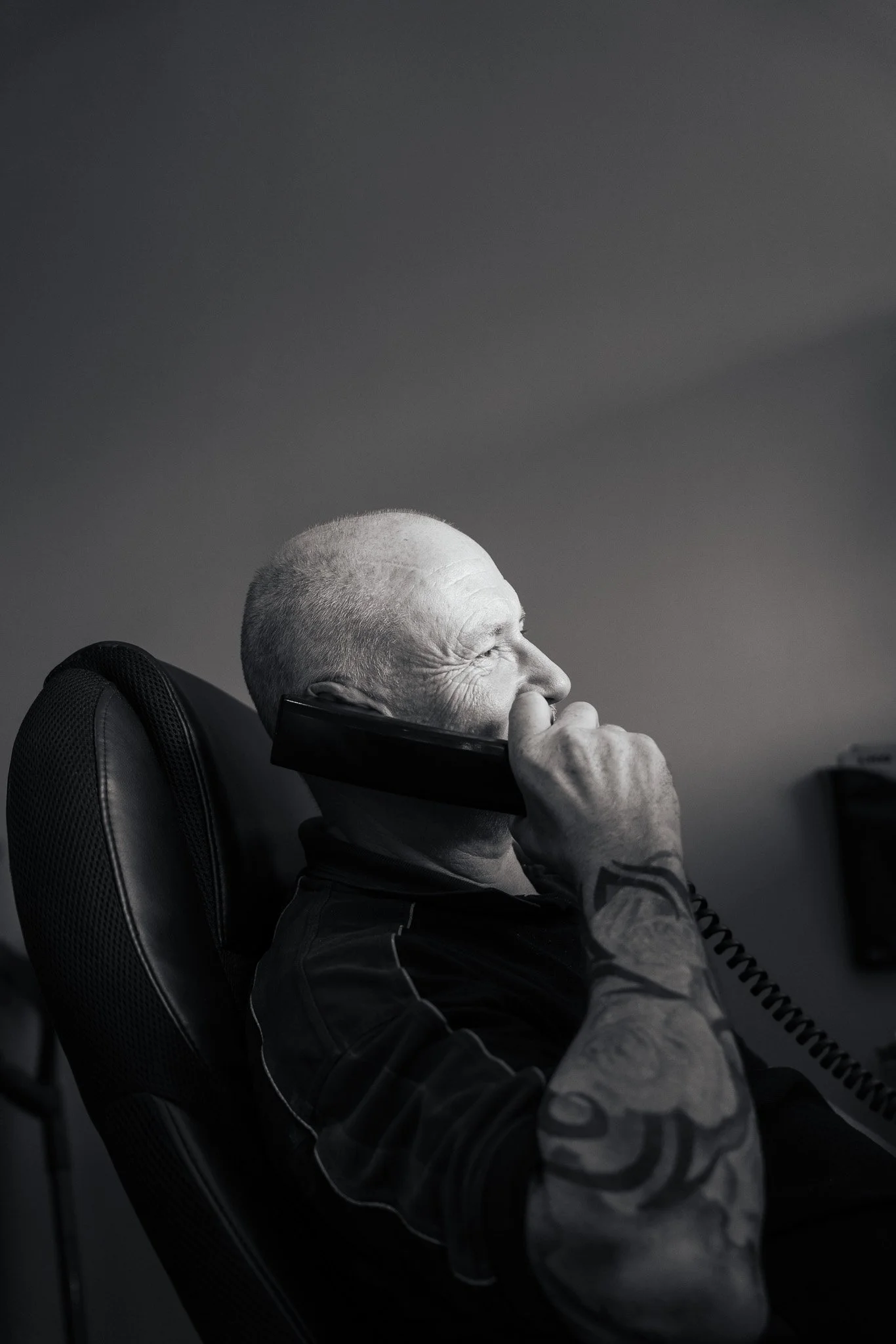 A black and white photo of an elderly man with a shaved head, sitting in a chair, talking on the phone, and smiling. He has tattoos on his arm.