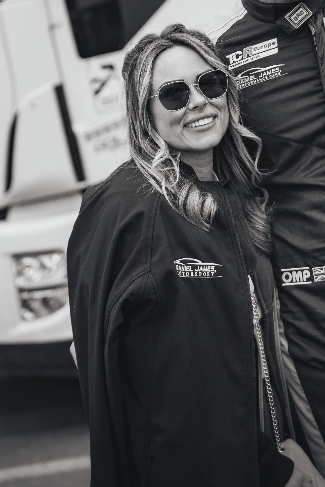 A woman smiling and wearing sunglasses, standing in front of a truck, dressed in a black racing jacket with sponsor logos.