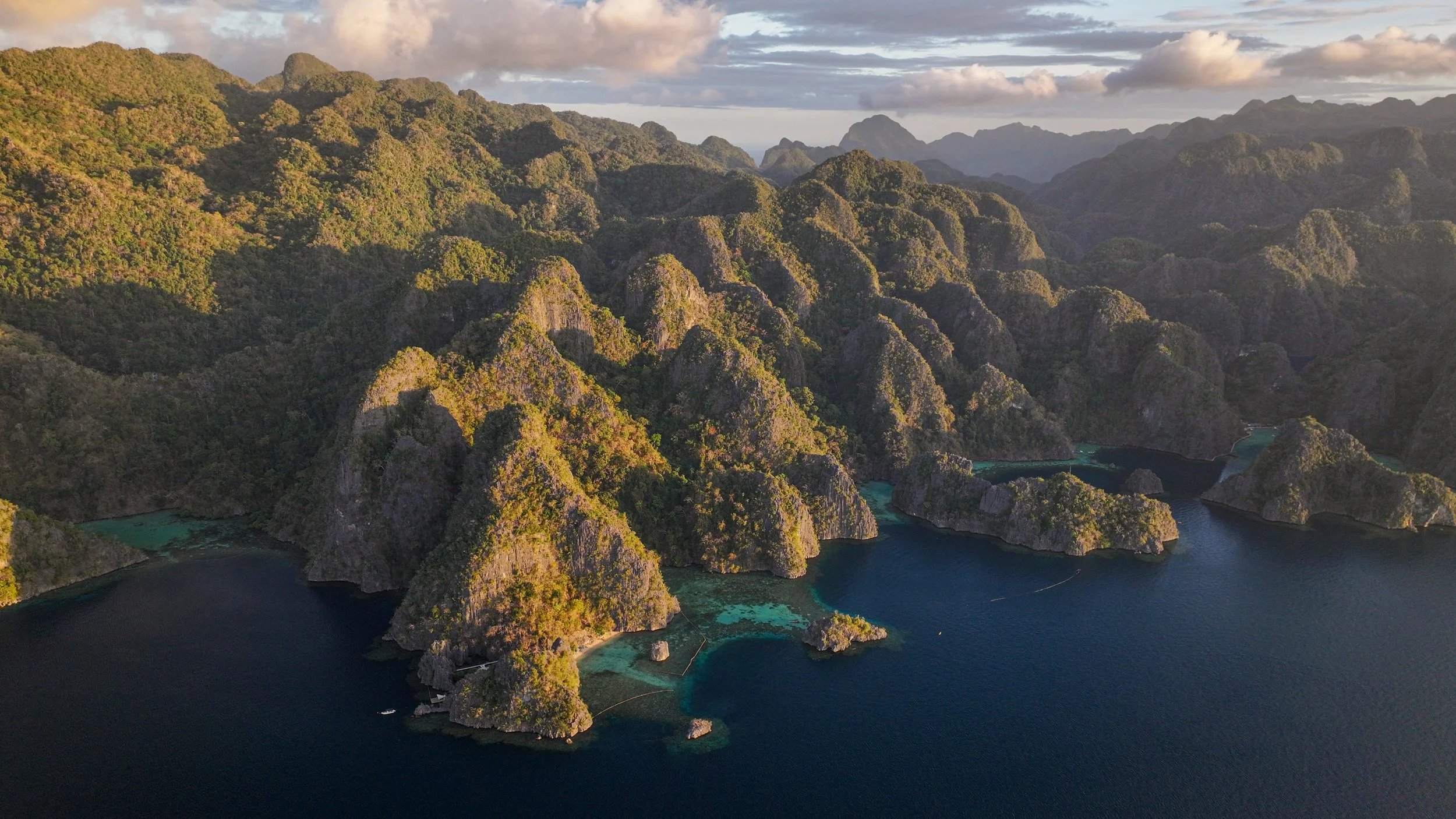 Aerial view of mountainous islands with lush greenery surrounded by deep blue water, featuring rugged limestone formations and patches of coral reefs, under a partly cloudy sky.